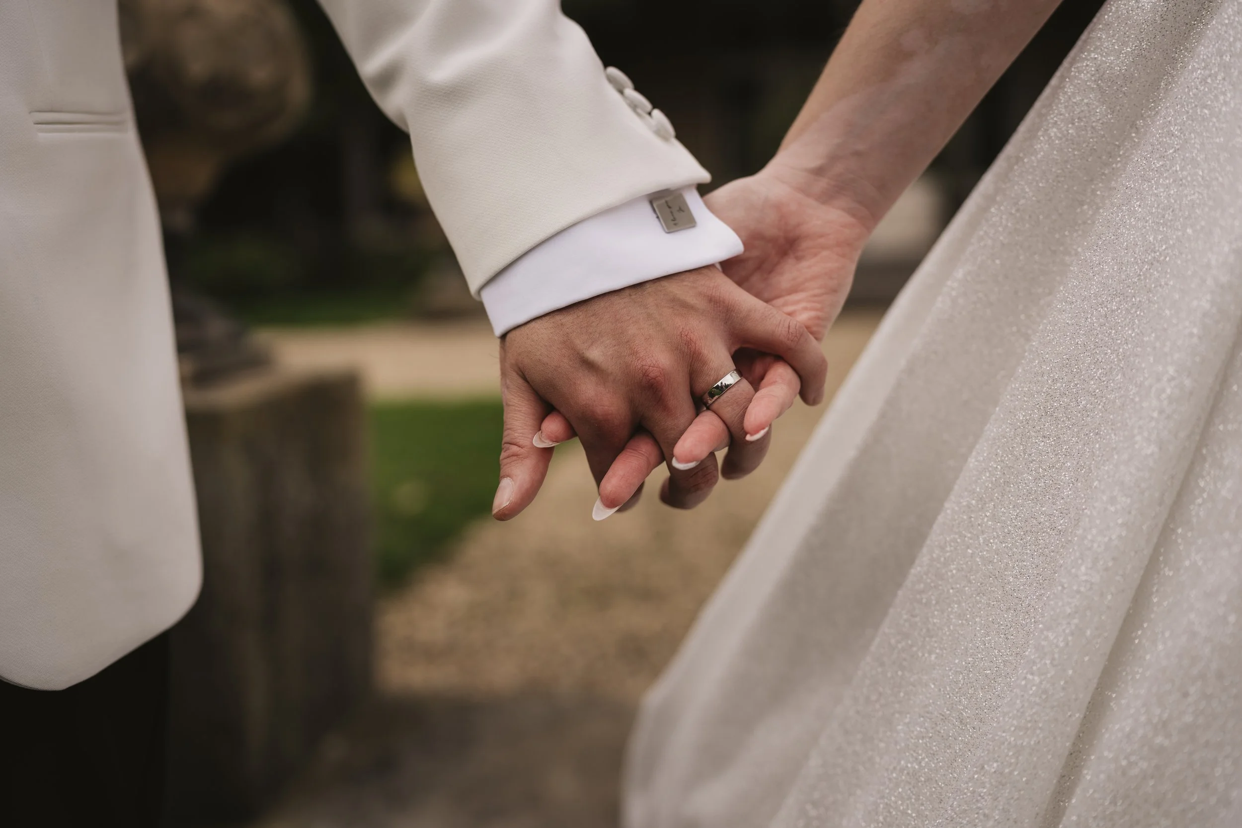 Close-up of a couple holding hands during their wedding, with the bride wearing a white dress and the groom in a cream tuxedo, outdoors.