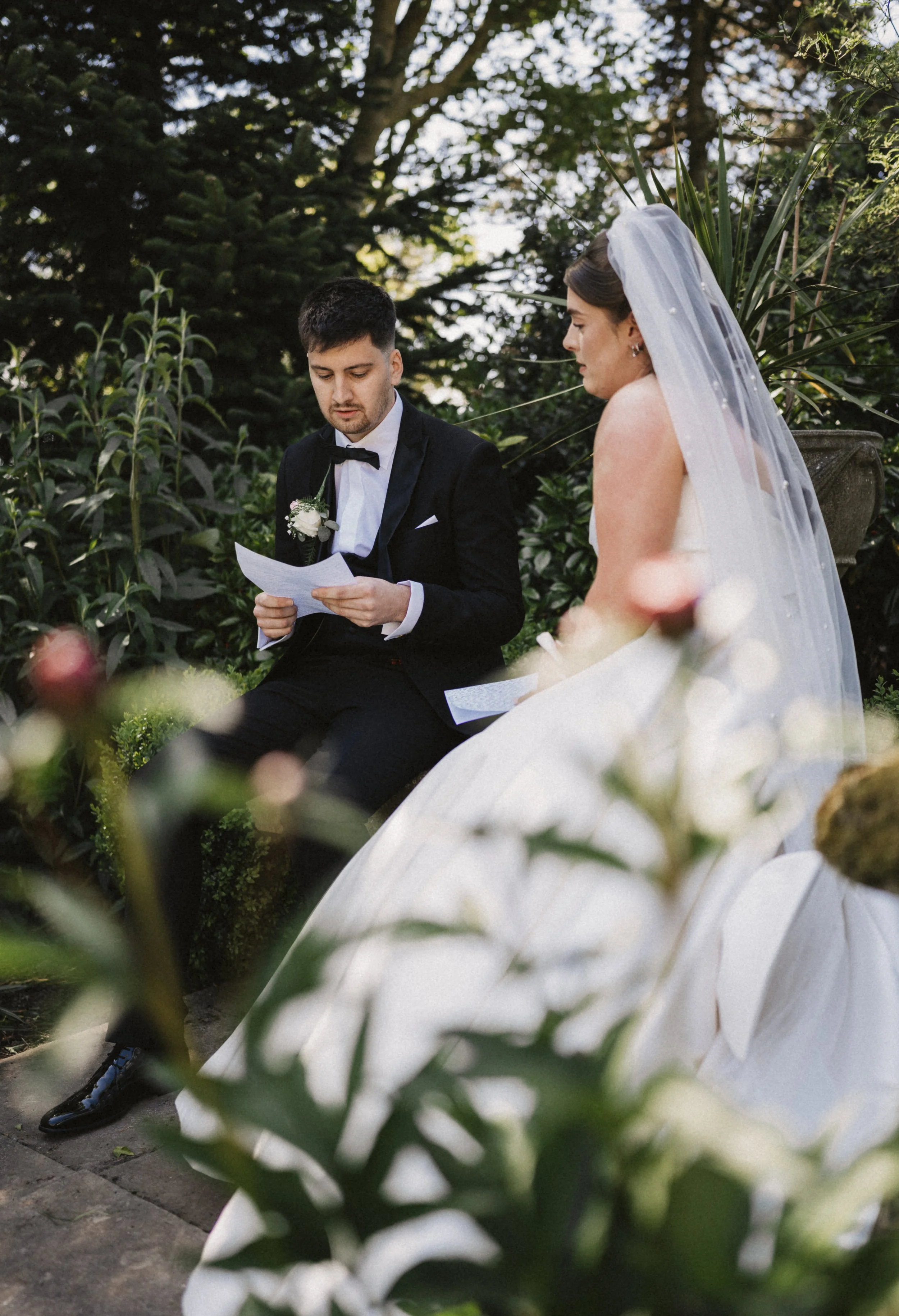 A bride and groom are sitting outdoors, with the groom reading a letter or vows. The bride is wearing a white wedding dress and veil, and the groom is dressed in a black tuxedo with a bow tie. There are lush green trees and plants in the background.