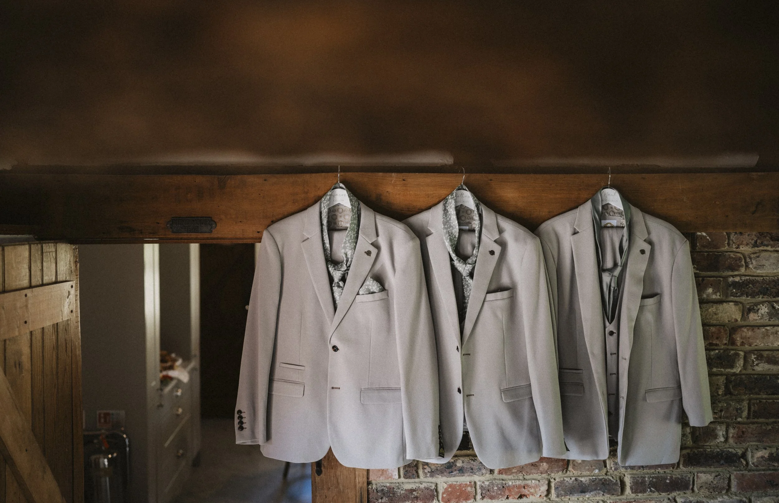 Three light gray suits hanging on a wooden beam against a brick wall.