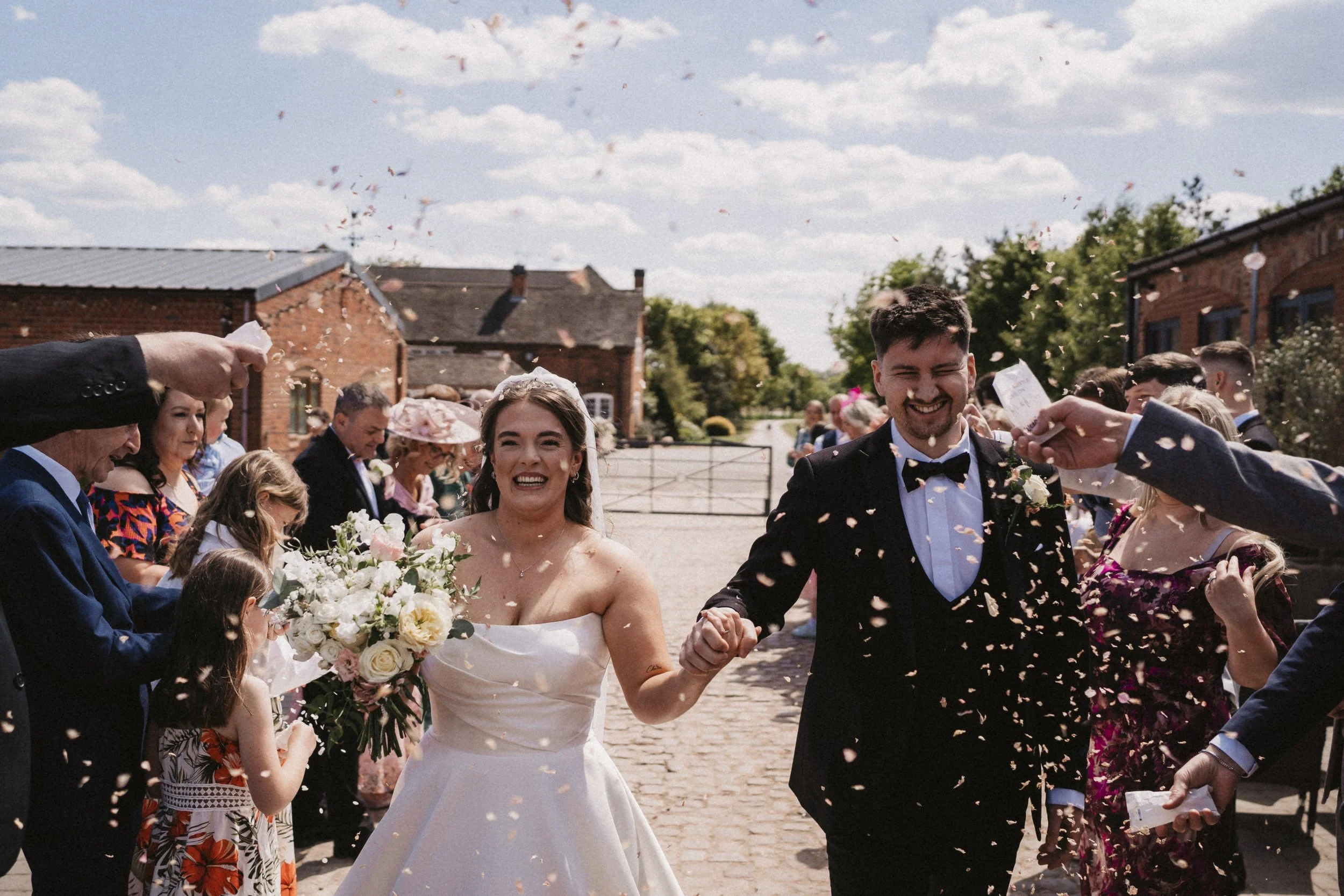 A newlywed couple walking hand-in-hand through a celebration of wedding guests throwing flower petals during daytime outside a rustic brick building.