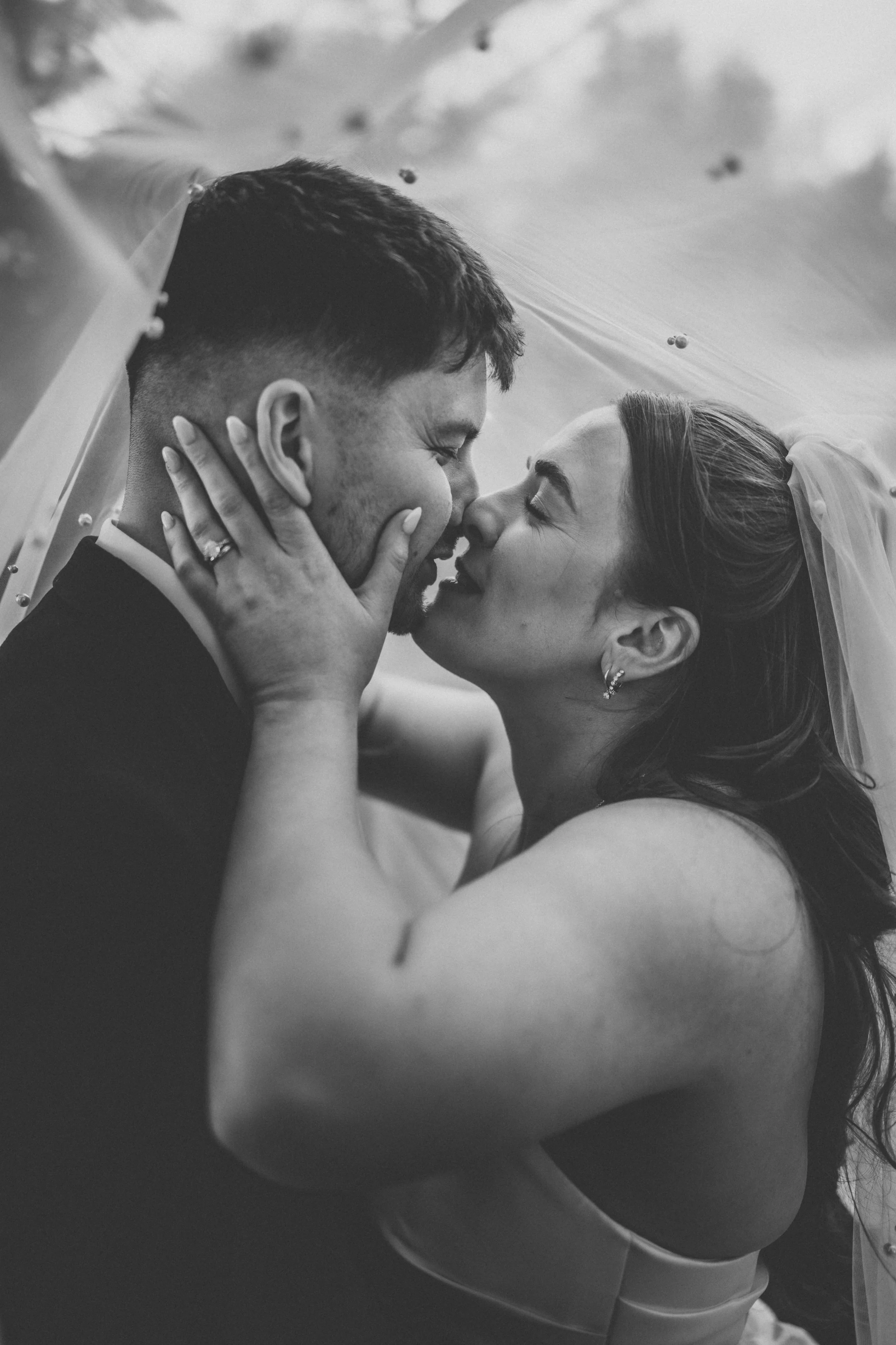 A black-and-white close-up of a newlywed couple sharing a kiss, with the bride's hand on the groom's face and her wedding veil visible.