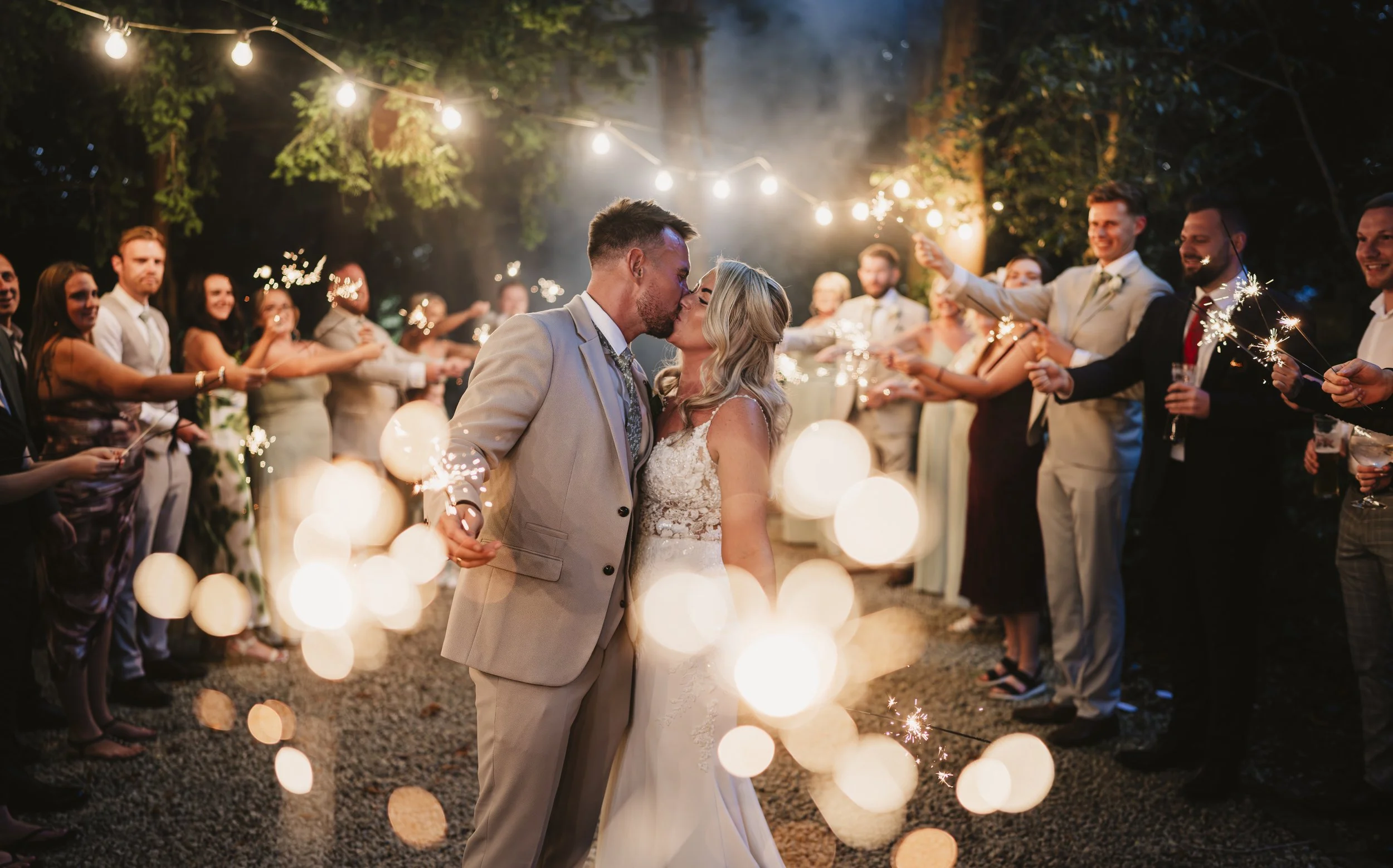A newlywed couple sharing a kiss at night, surrounded by friends holding sparklers in a garden celebration.