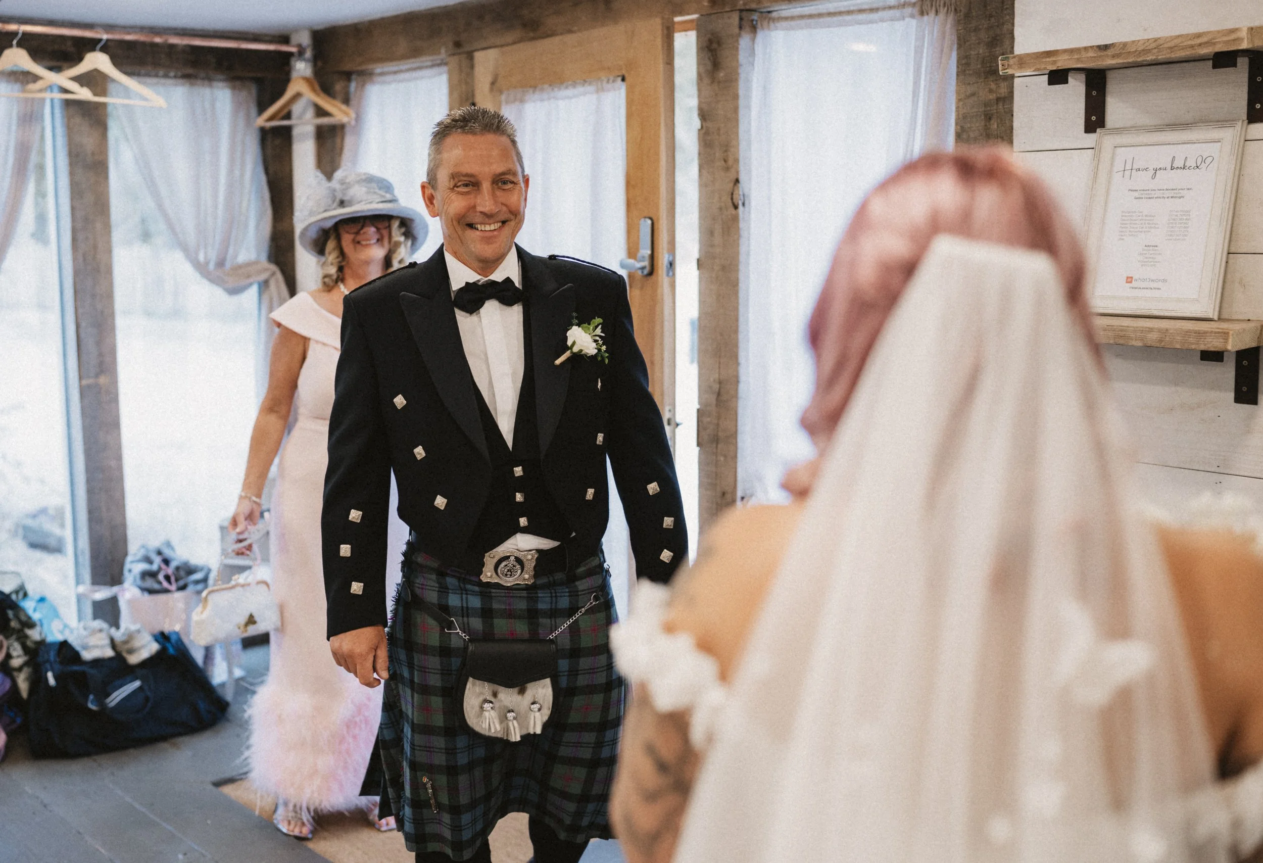 A groom in a traditional black tuxedo and Scottish kilt smiling at a bride with a veil, inside a rustic wedding venue with wooden walls and large windows.