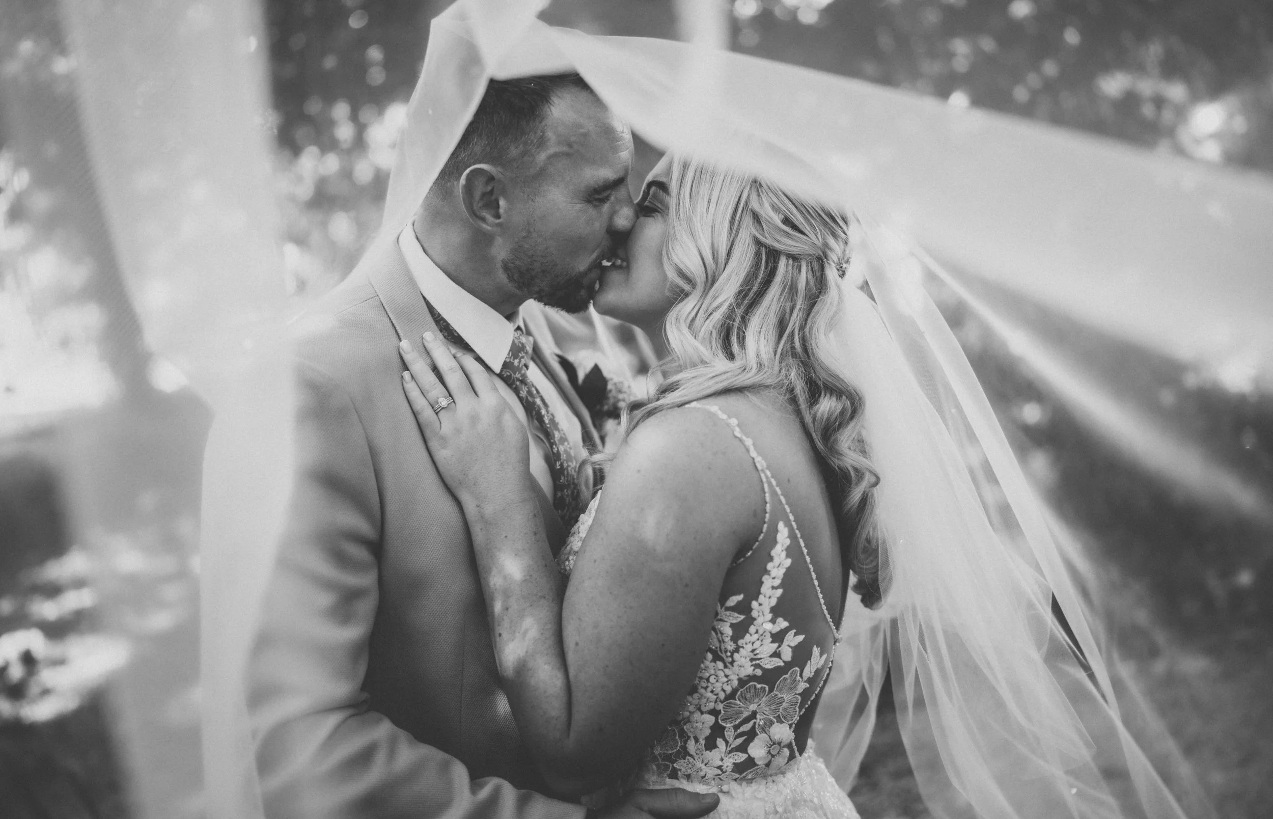 Black-and-white photo of a newlywed couple under a wedding veil, embracing and kissing outdoors, with trees blurred in the background.
