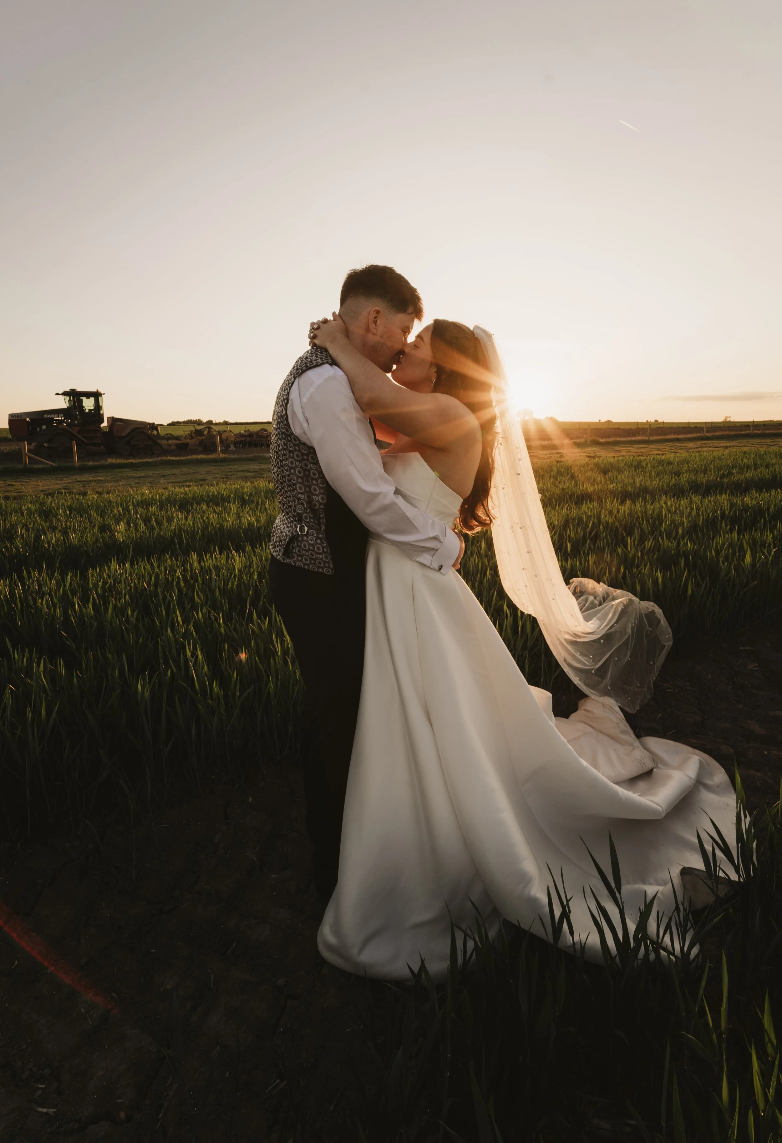 A bride and groom kissing in a field at sunset, with the bride wearing a white wedding gown and veil, and the groom in a shirt and vest, with farm equipment in the background.