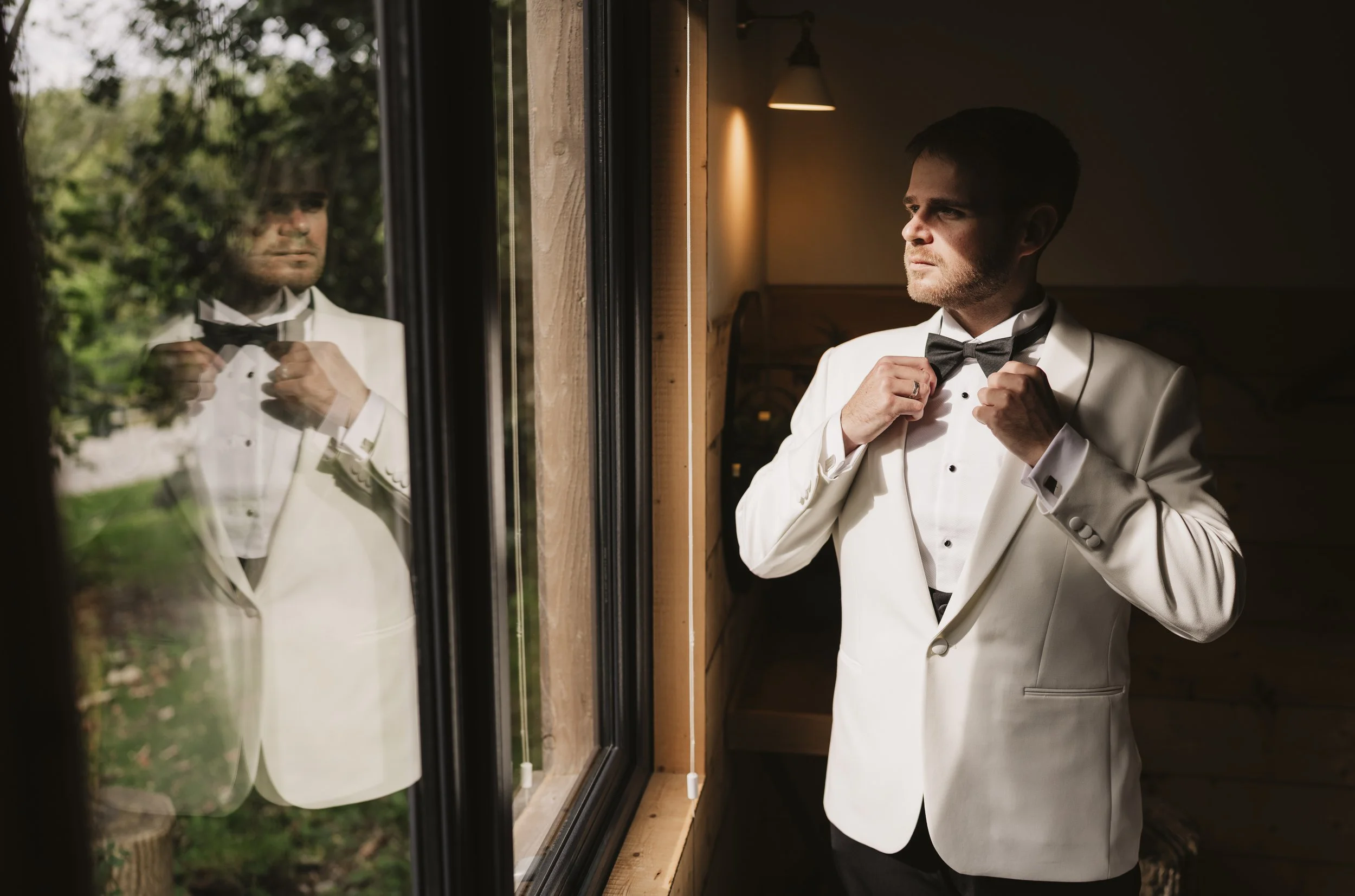 A man in a tuxedo adjusting his bow tie while looking out of a window. His reflection is visible on the glass.