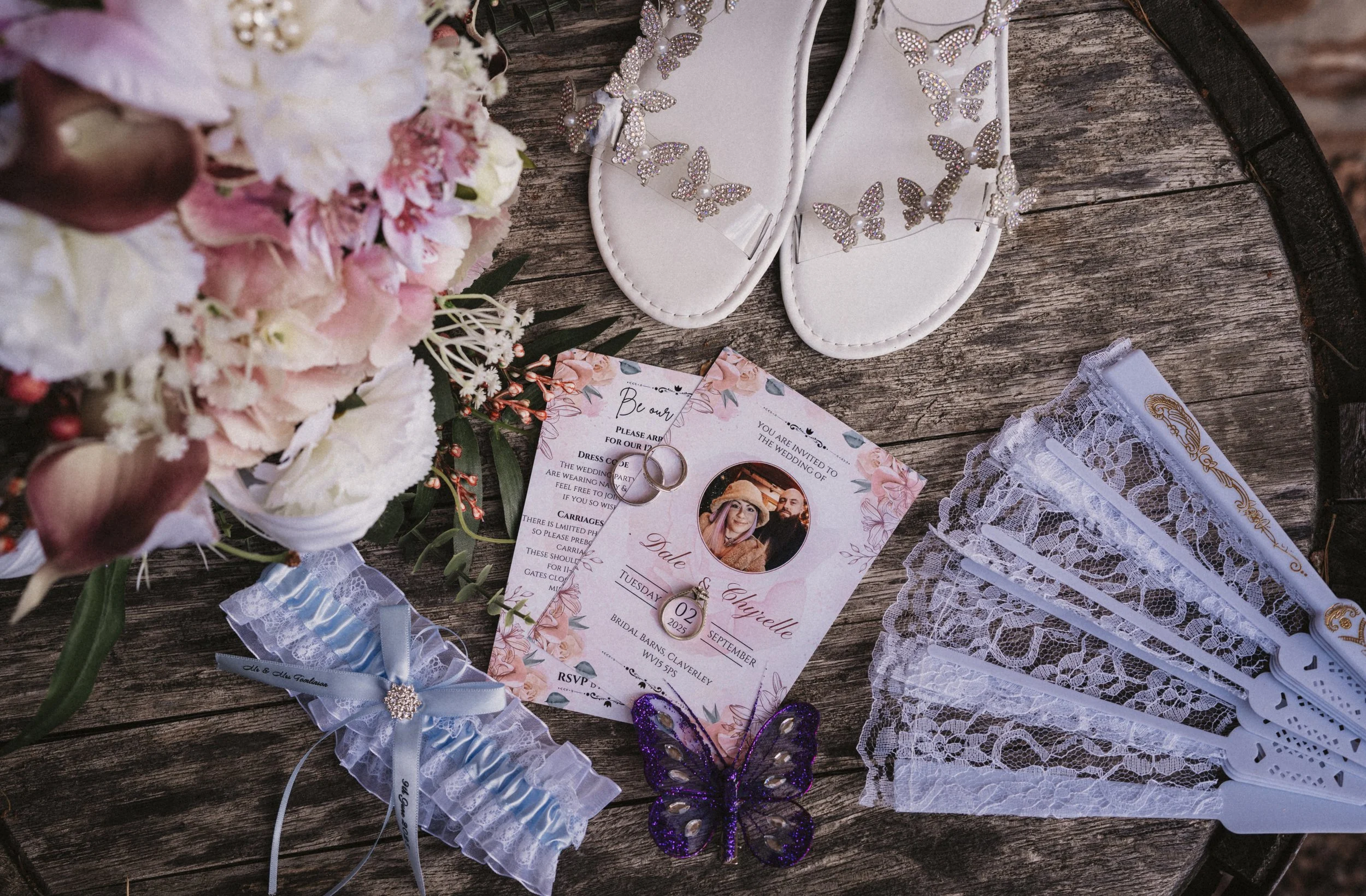 Wedding invitation, rings, lace garter, butterfly decoration, white sandals with butterfly embellishments, lace fans, and a flower bouquet on a rustic wooden table.