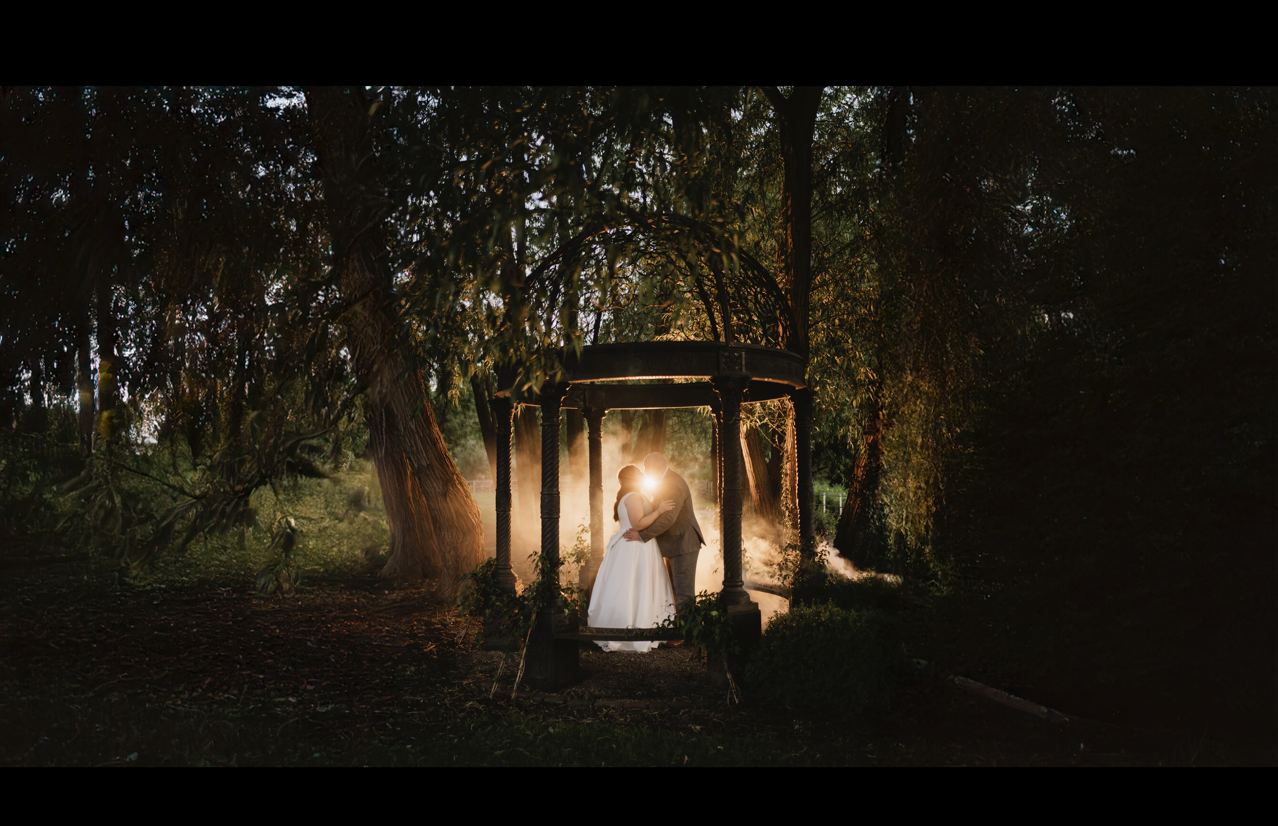A newlywed couple sharing a kiss during sunset under a gazebo in a forested area surrounded by trees and mist.
