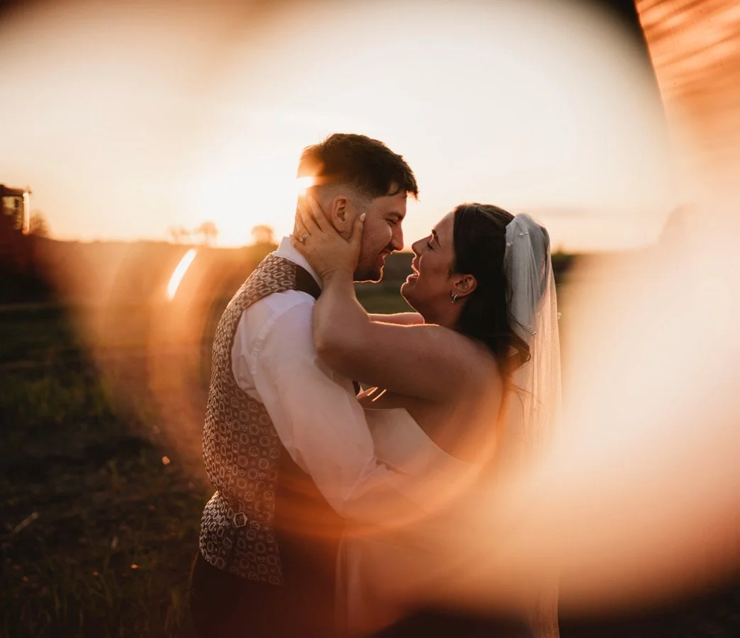 A bride and groom embracing at sunset, with the sun backlighting them and creating a warm glow around their faces.