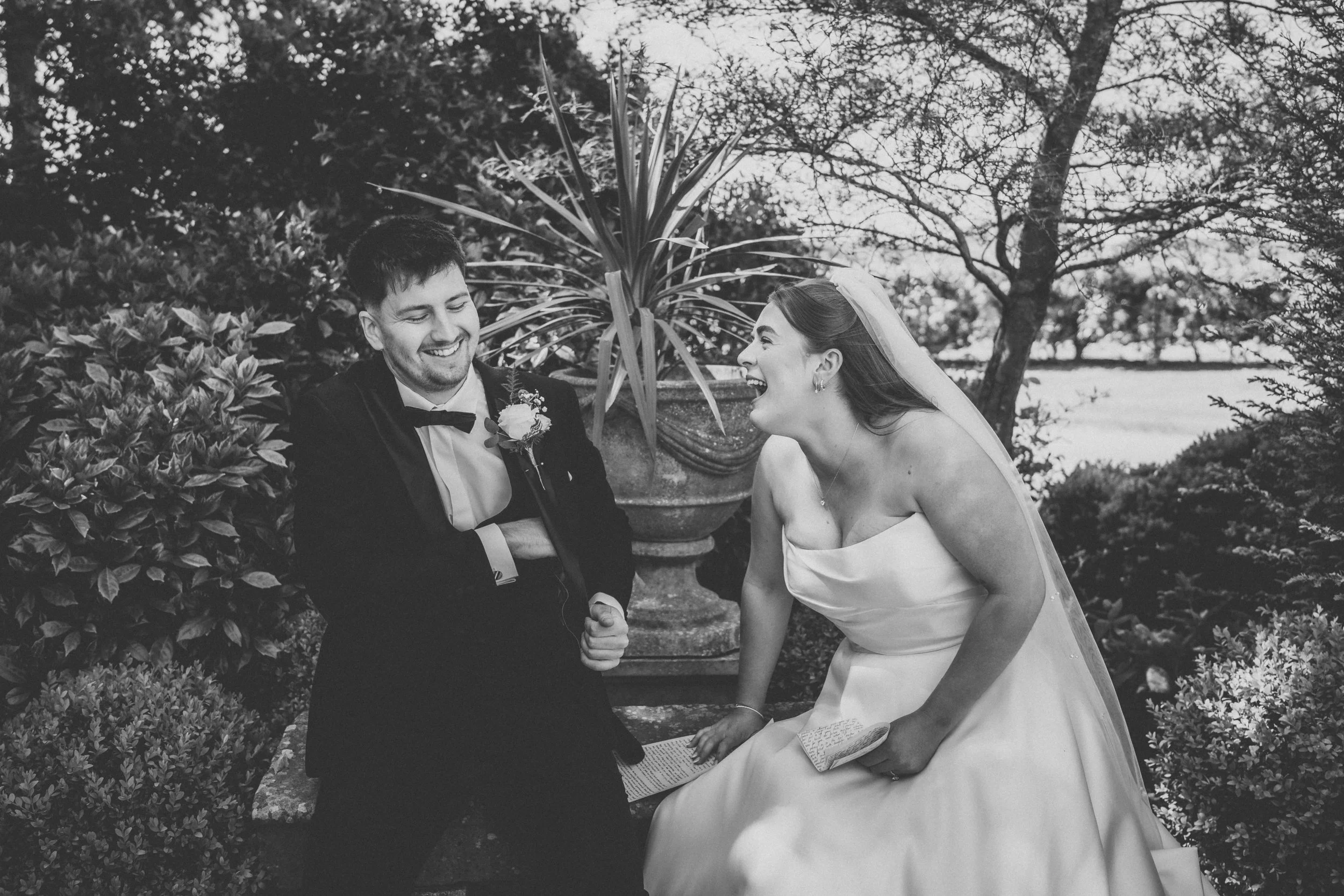 A black and white photograph of a bride and groom sitting outdoors, laughing together. The bride is wearing a strapless wedding gown and veil, while the groom is dressed in a tuxedo with a bow tie. They are surrounded by bushes and trees.