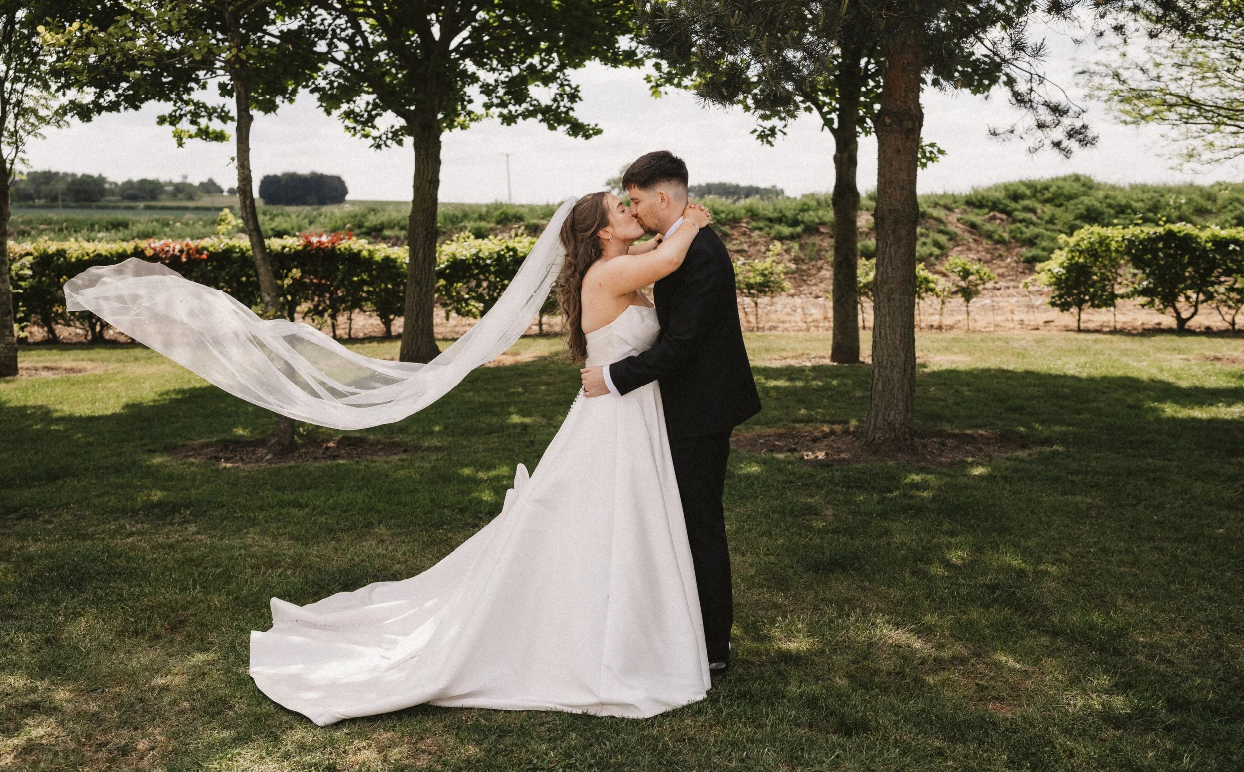 A bride and groom kissing outdoors on a grassy area with trees and shrubs in the background, the bride's veil flowing in the wind.