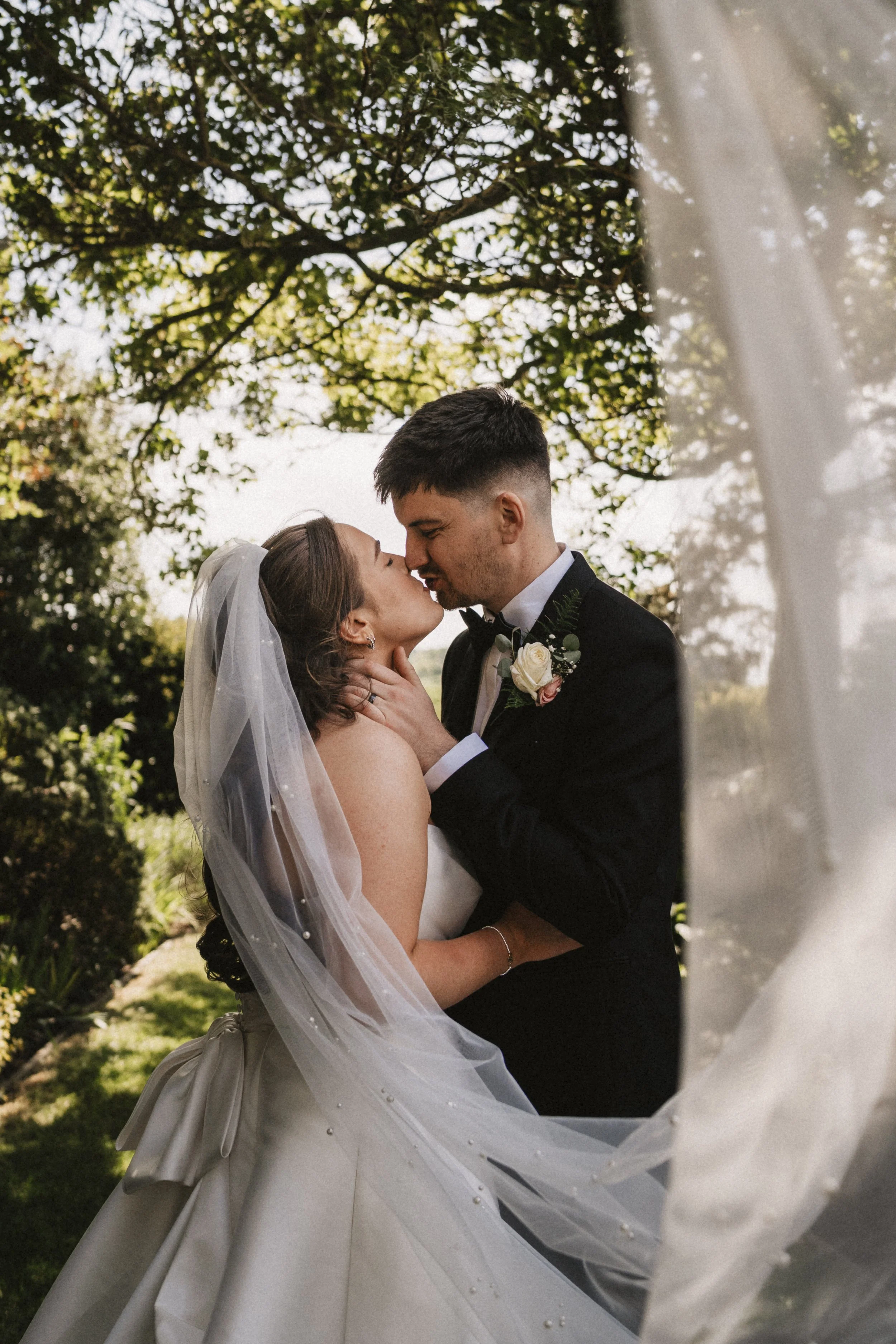 Bride and groom sharing a kiss outdoors, framed by a white veil, with trees and sky in the background.