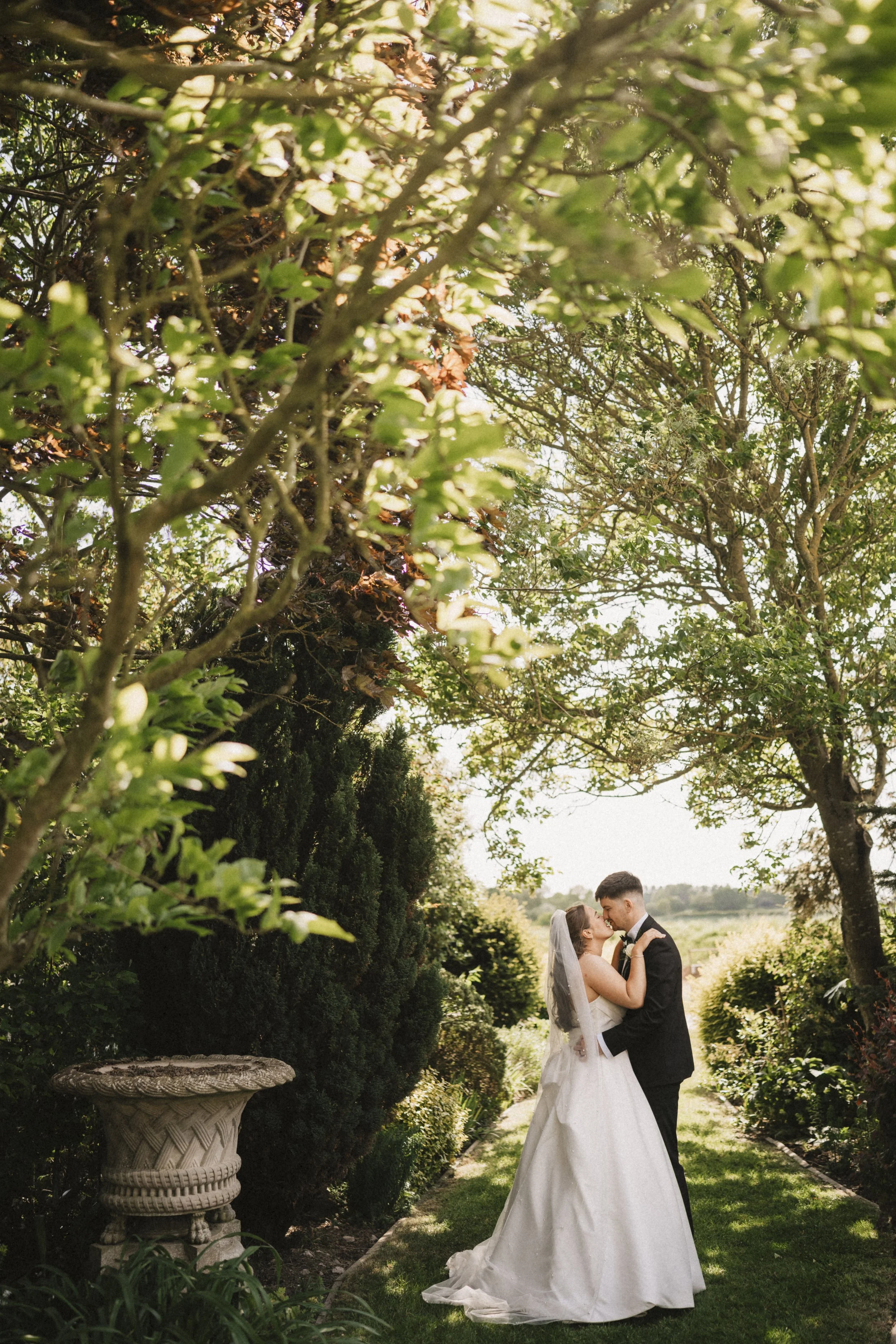 A bride and groom embracing and kissing in a lush garden with green trees and bushes on a sunny day.