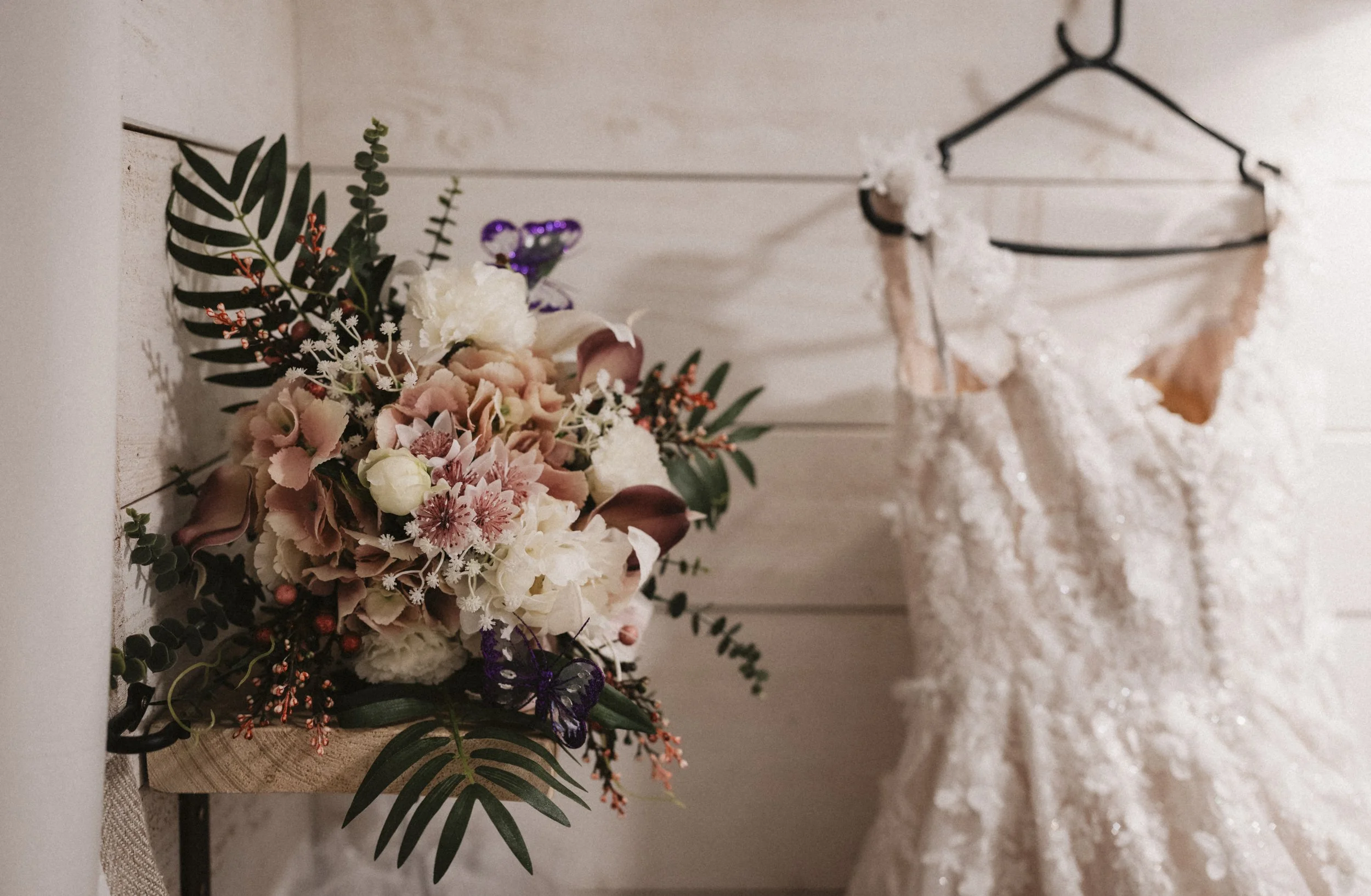 A wedding bouquet with pink, white, and burgundy flowers, green leaves, and decorative elements placed on a wooden surface. In the background, a lace wedding dress hangs on a black hanger against a white wall.