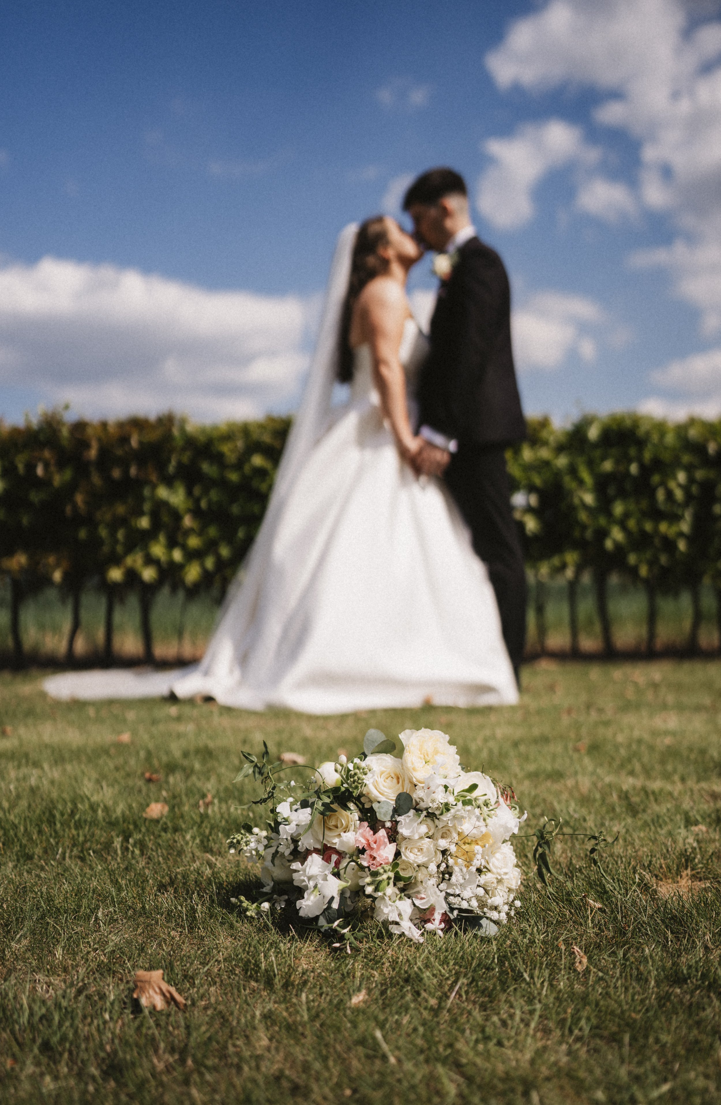 Wedding bouquet with white and pink flowers on grass, blurred bride and groom kissing in background against blue sky.