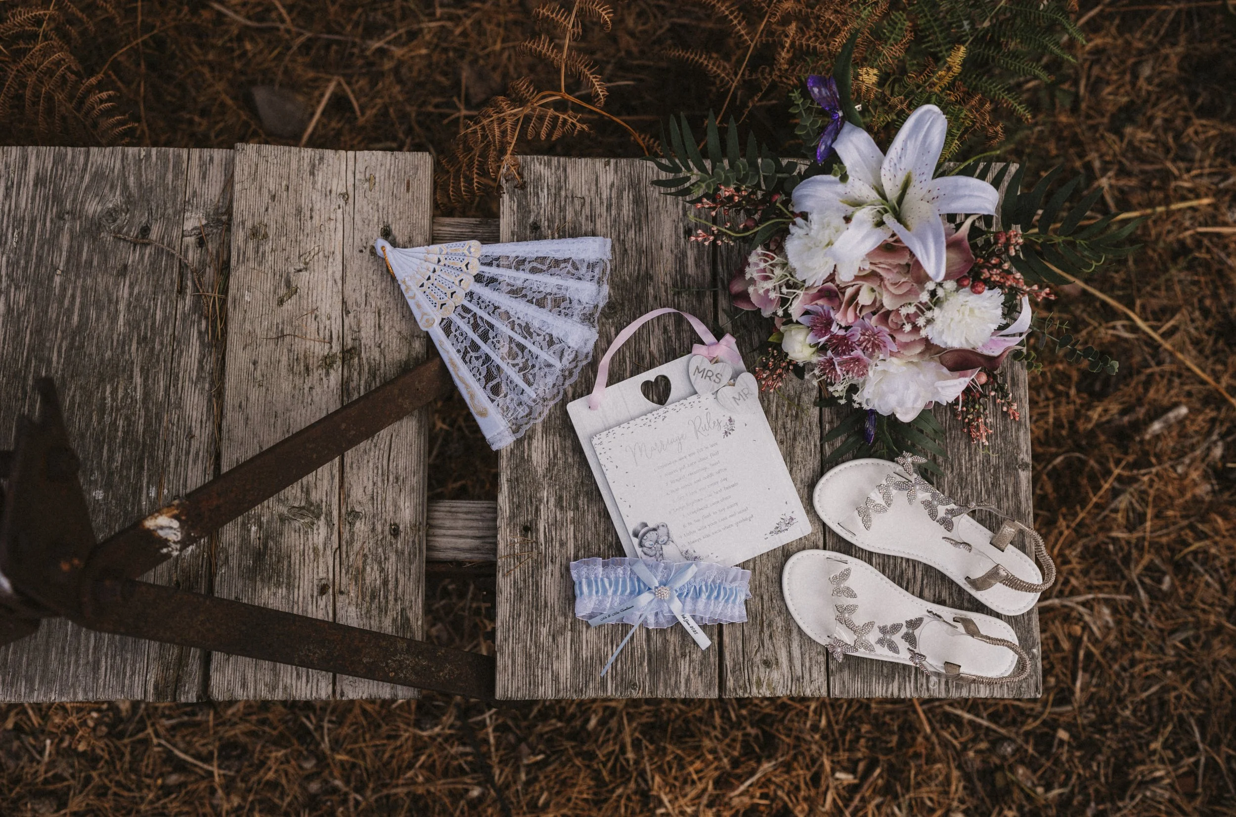Wedding accessories including white sandals with gold accents, a lace garter, a wedding playlist or program, a floral bouquet with white lilies, pink, purple flowers, and greenery, and a small lace-detailed fan on a rustic wooden table outside.