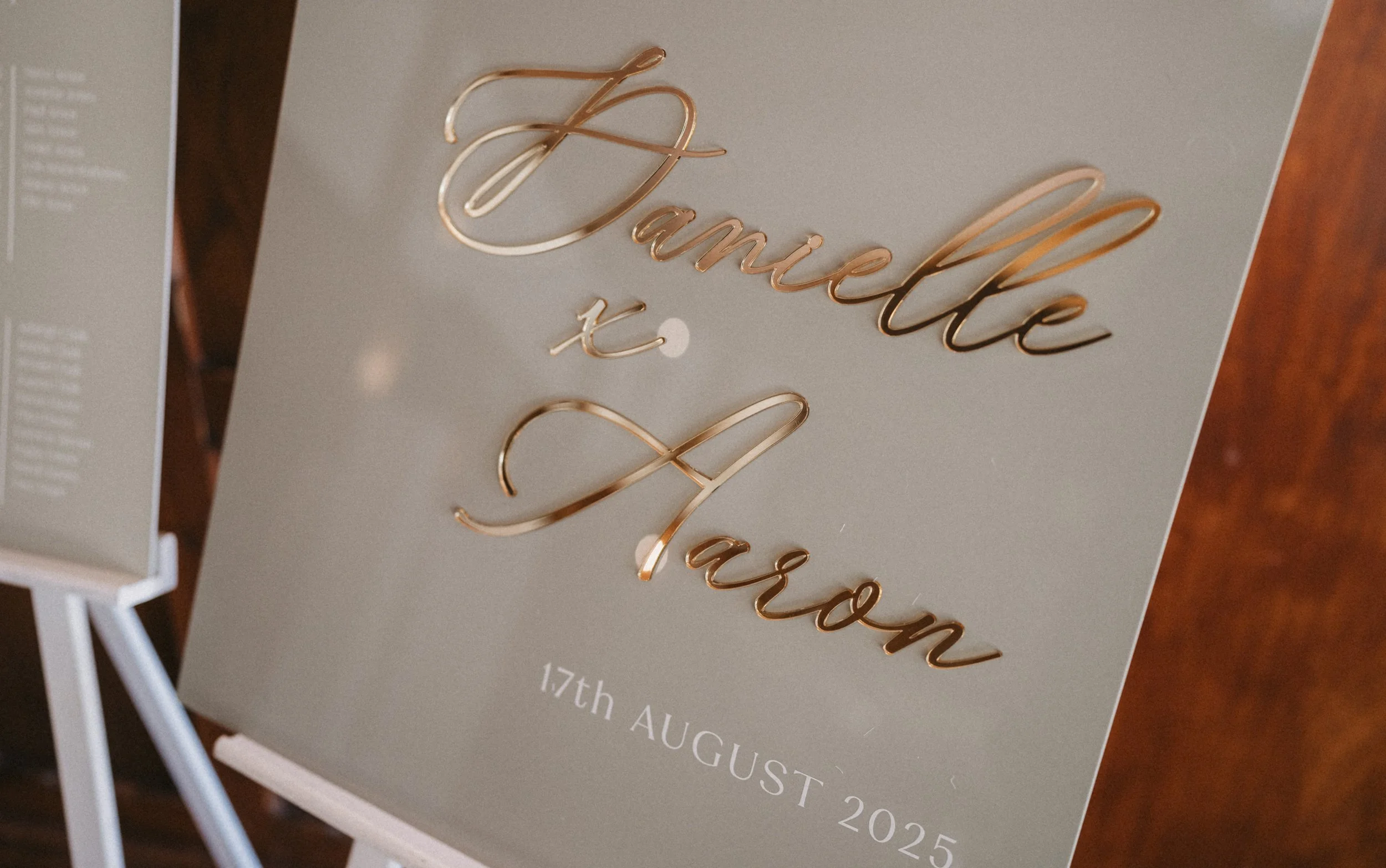 A wedding sign with gold metallic cursive text reading "Danielle x Jason" and the date "17th August 2025" below, on a light-colored background.
