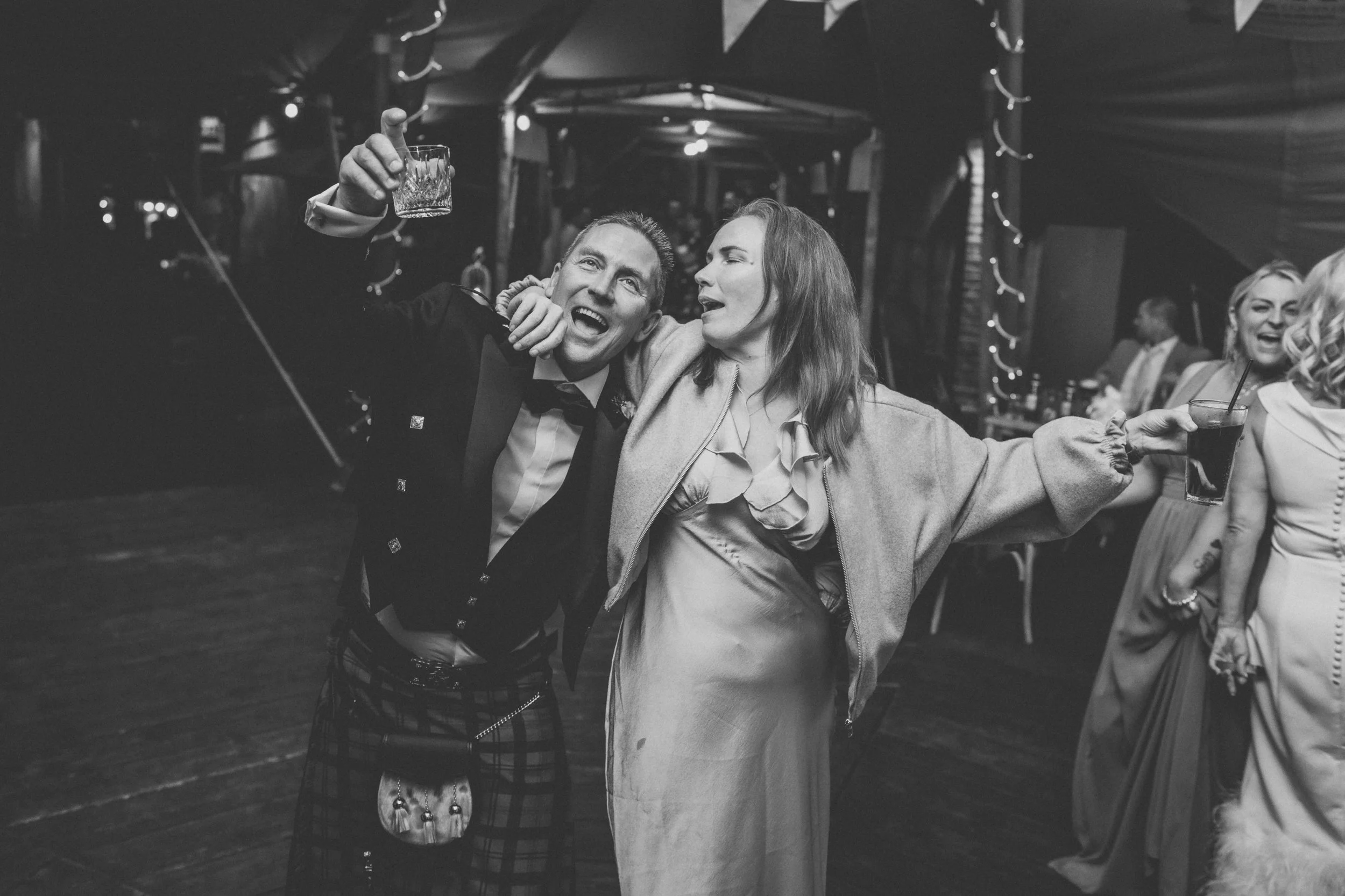 Black and white photo of a man wearing a tuxedo and a woman in a dress, dancing and holding drinks at a party or celebration, with other people in the background.