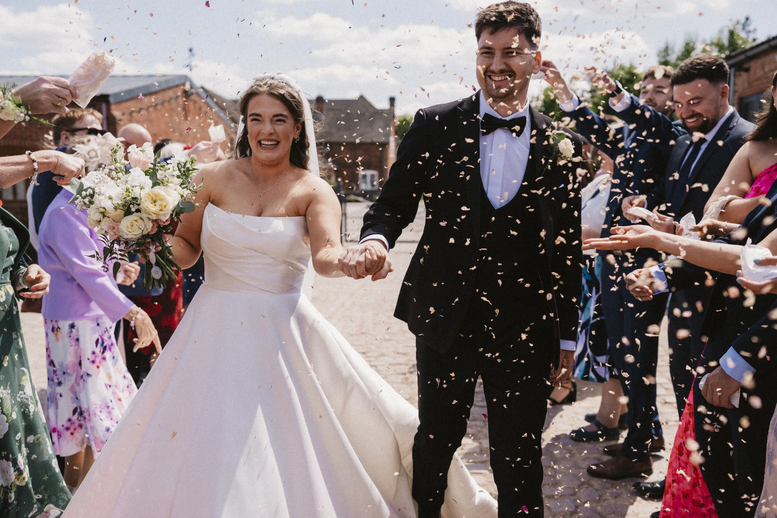 A newly married couple on their wedding day, smiling and holding hands, surrounded by guests throwing confetti outdoors.