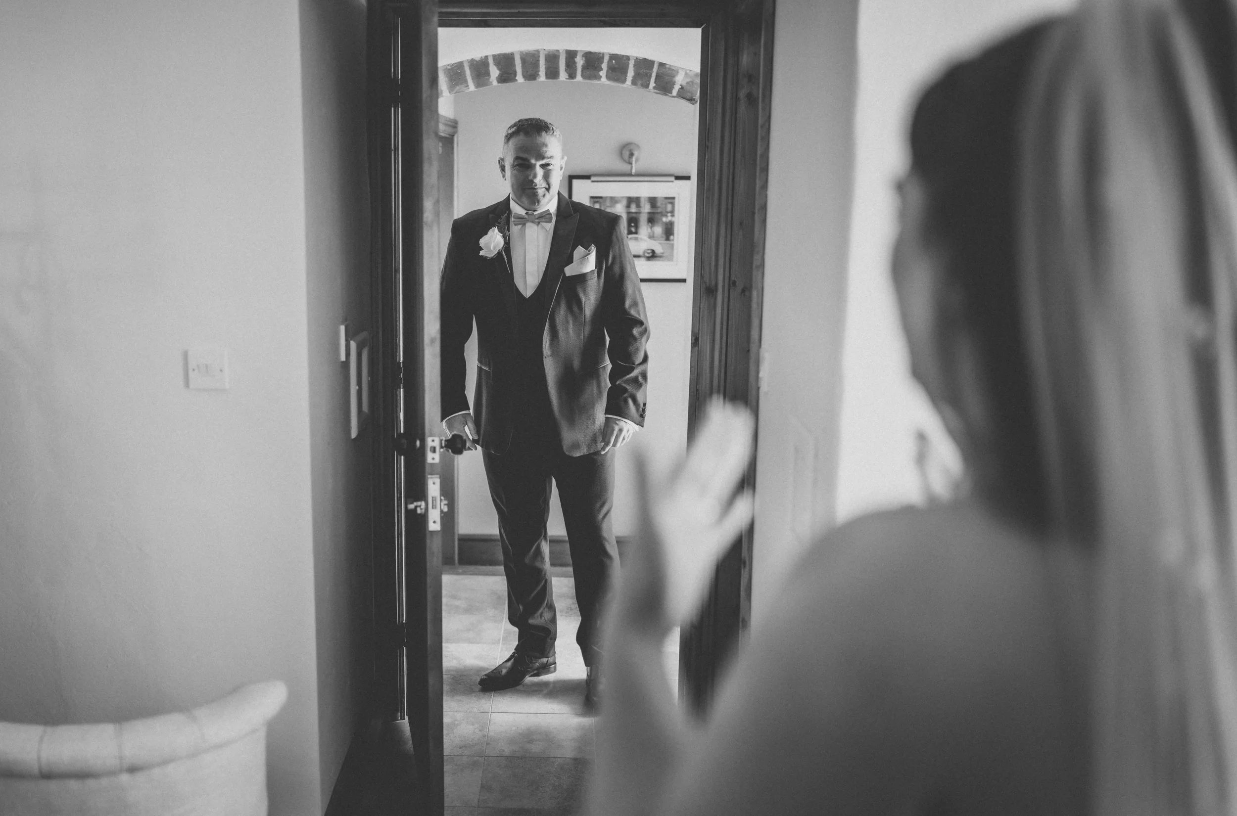 A groom in a tuxedo standing in a doorway, looking at a bride who is partially visible and waving.
