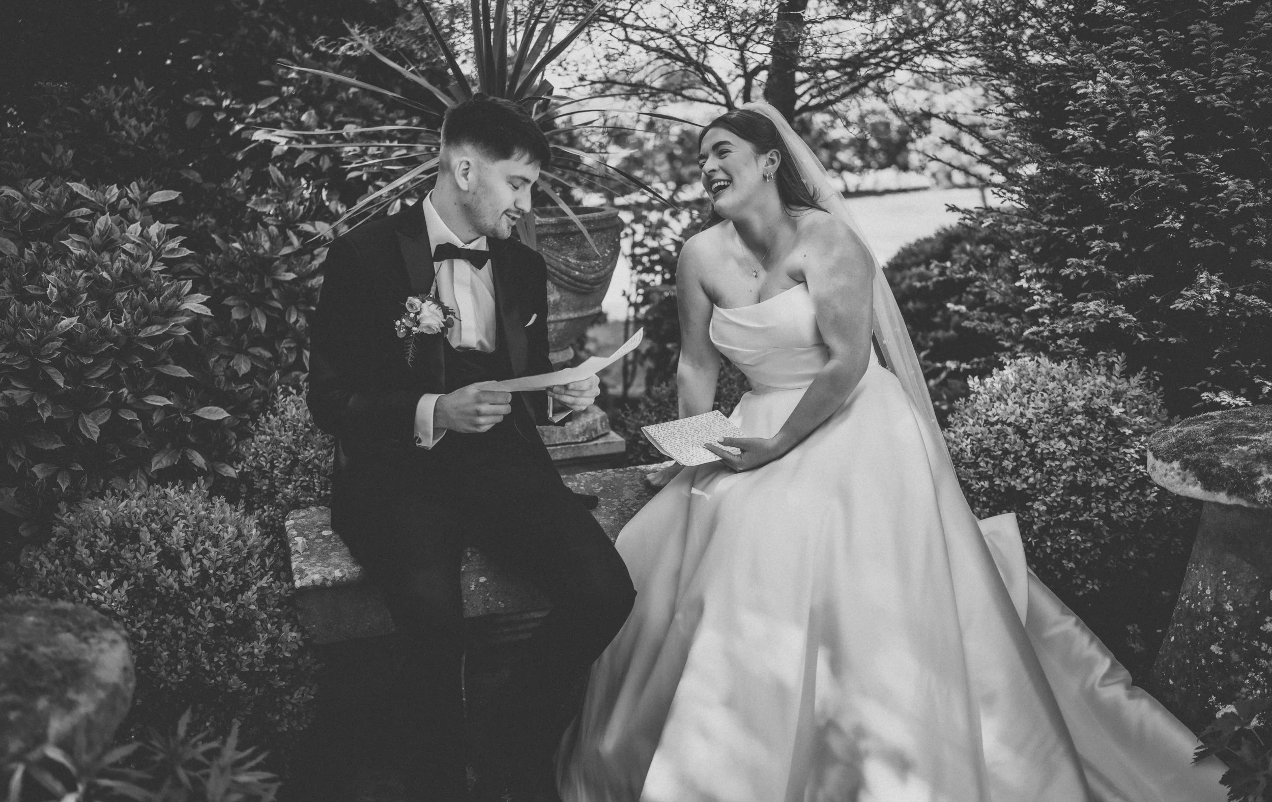 A black and white photo of a bride and groom sitting on a garden bench, exchanging vows or reading their vows, surrounded by plants, with trees in the background.