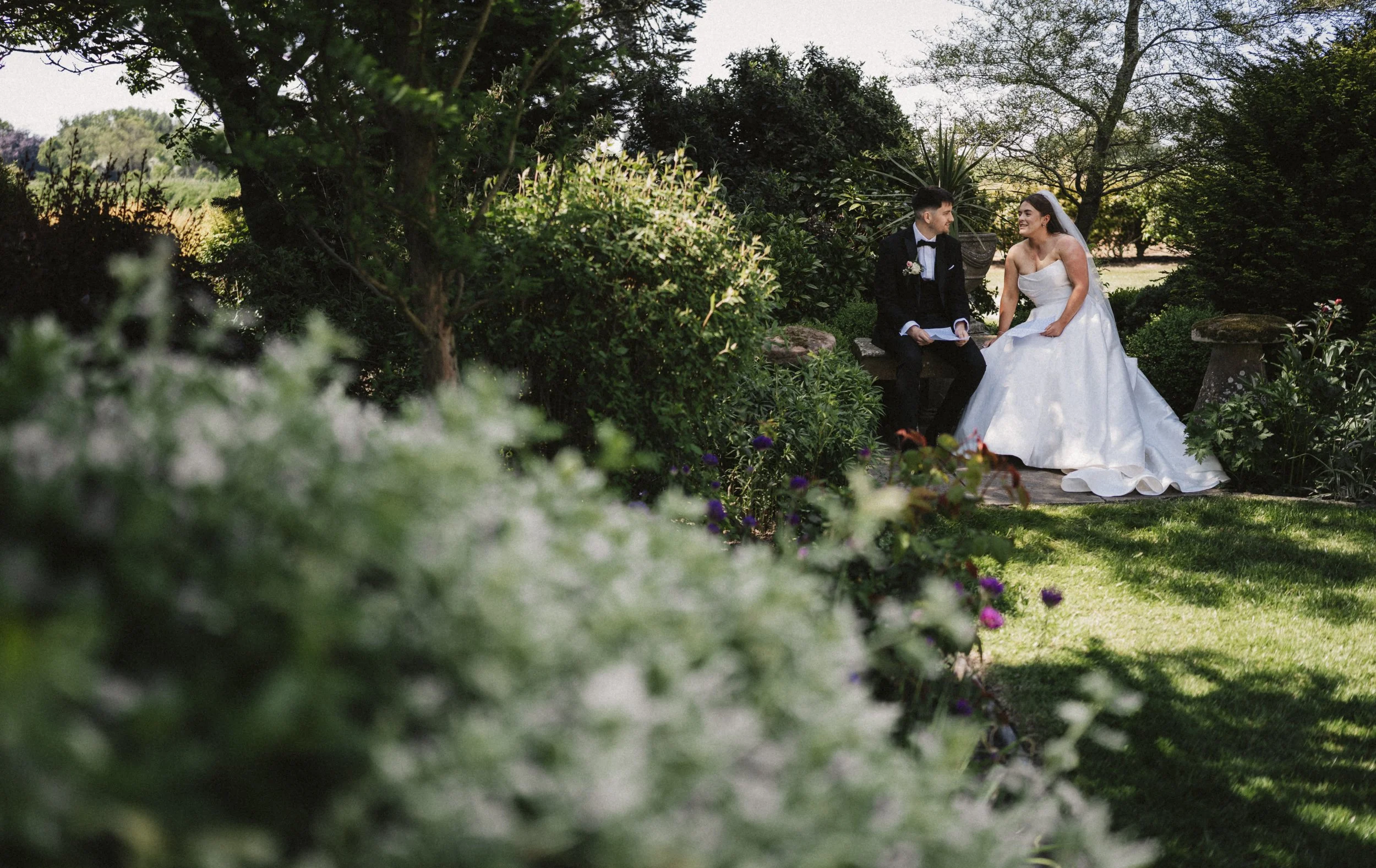 A bride and groom sitting on a stone bench in a garden during their wedding ceremony. The bride is in a white wedding gown and veil, and the groom is in a black tuxedo. They are looking at each other, surrounded by greenery and flowering plants.