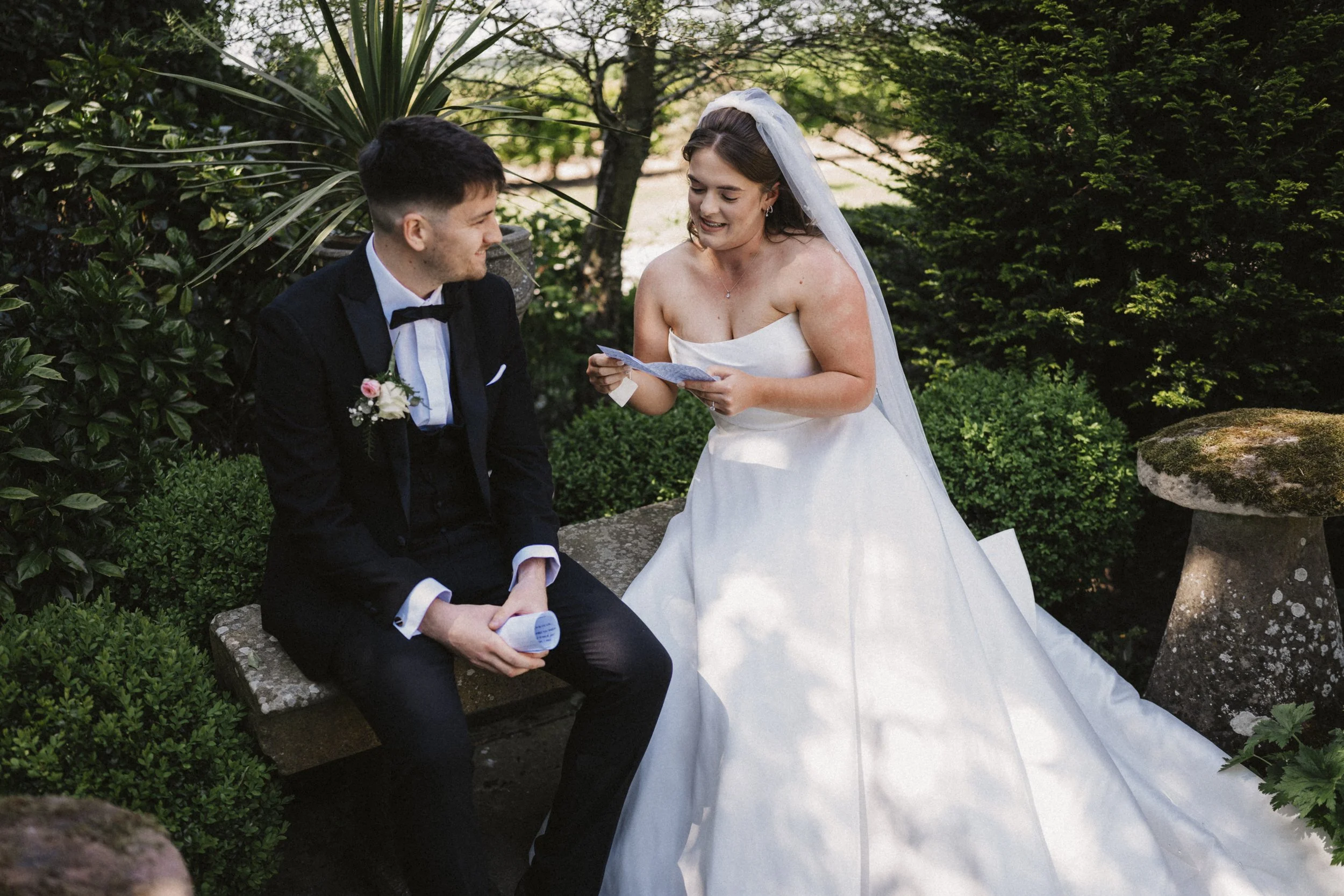 A bride and groom sitting on a bench outdoors during a wedding ceremony, with the bride reading vows or a letter, surrounded by green foliage.