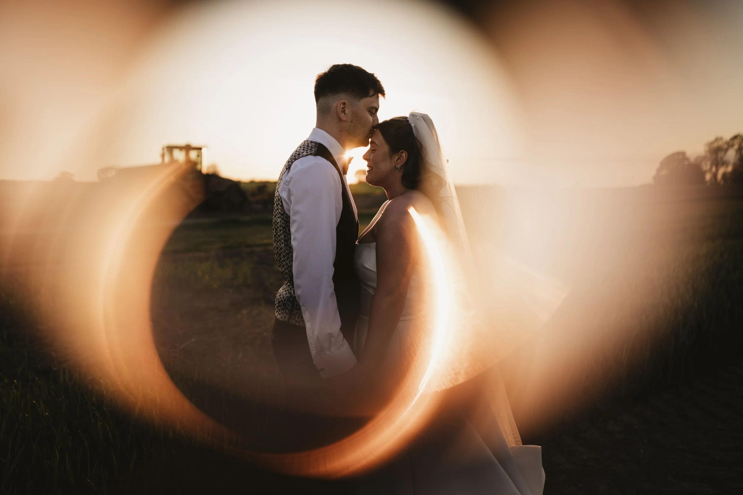 A bride and groom standing close together outdoors during sunset, sharing a tender moment with a lens flare creating a crescent shape around them.