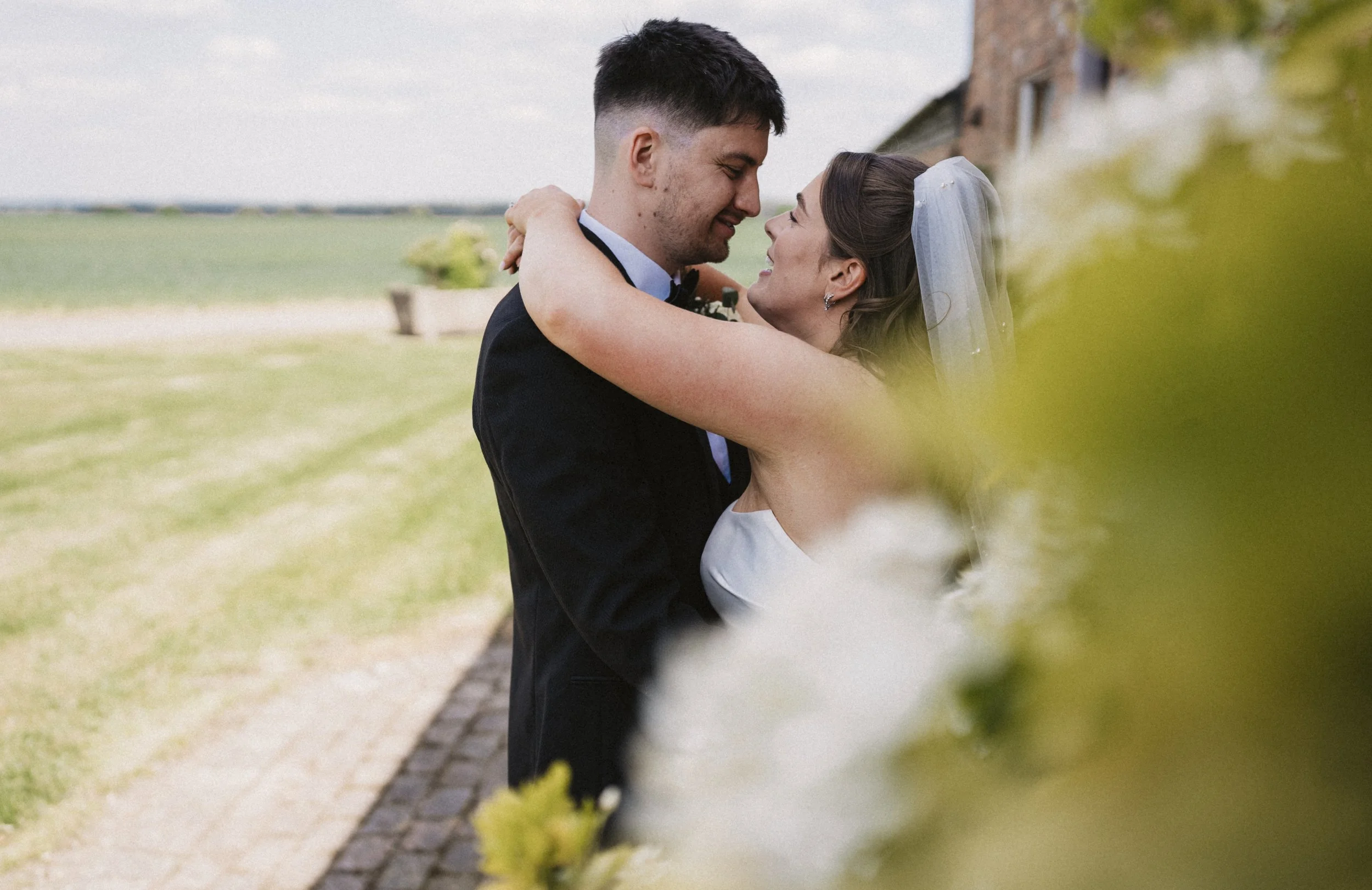 A bride and groom embrace outdoors on their wedding day, with the bride wearing a veil and a white dress, and the groom in a black tuxedo. They are smiling and close to each other amid a garden setting with a brick path and green landscape in the bac