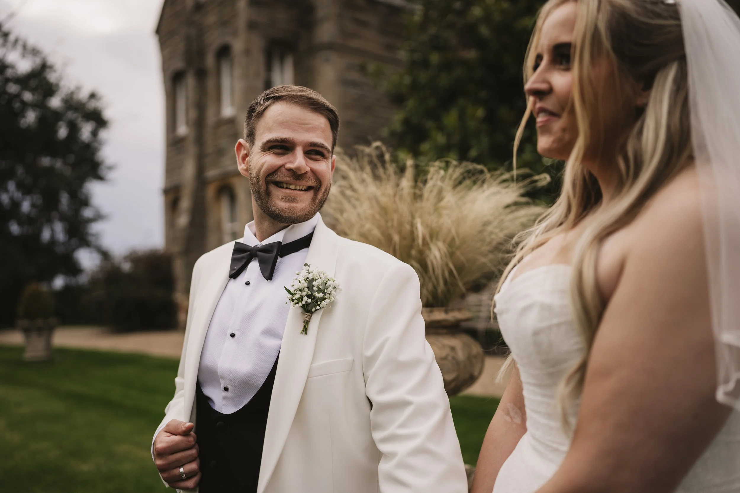 A groom in a white tuxedo with a black bow tie and boutonniere looks at a bride in a white wedding dress with a veil, outdoors near a historic building and greenery.