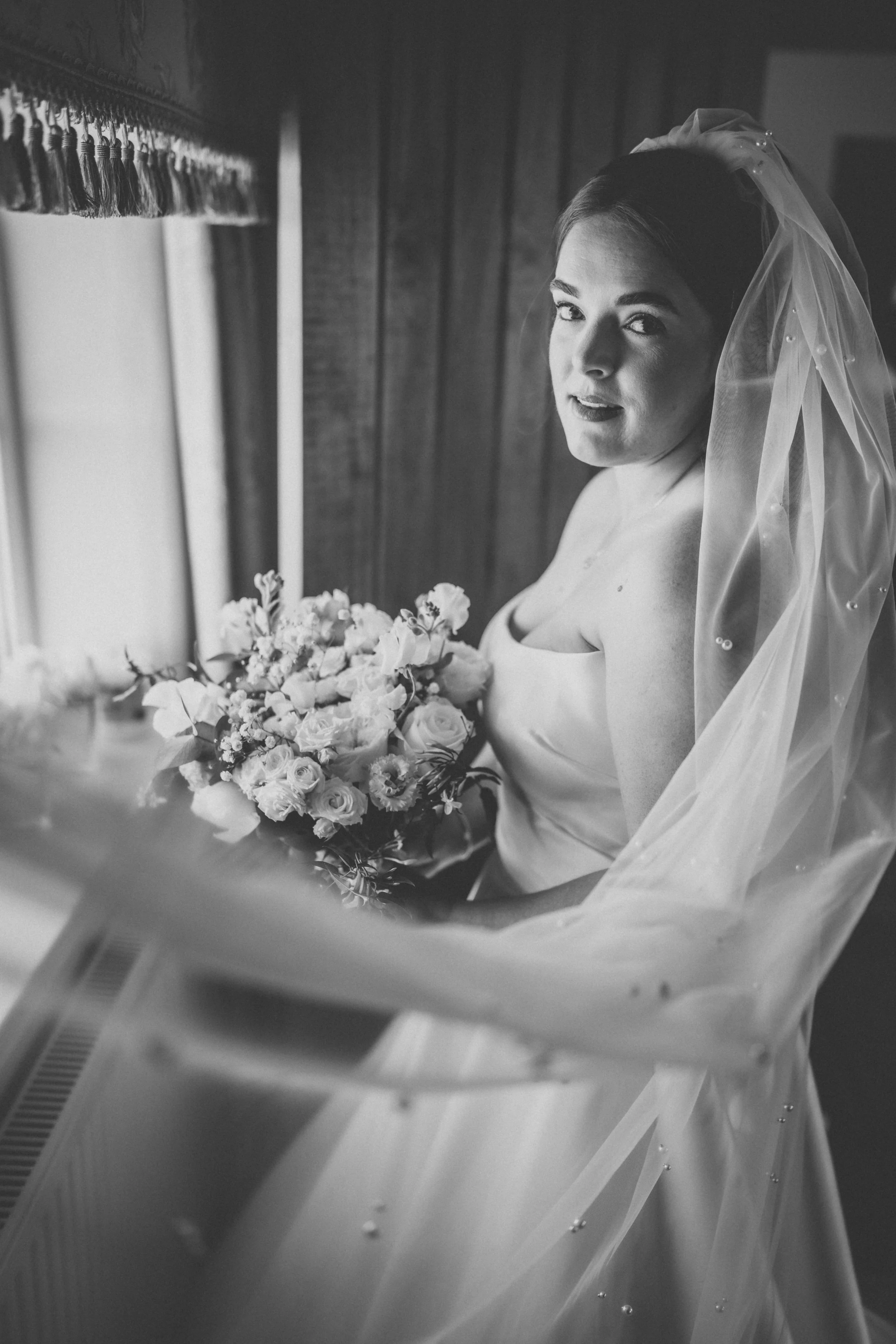 Black and white photo of a bride holding a bouquet, looking at the camera, wearing a wedding dress and veil, standing near a window with wooden paneling in the background.