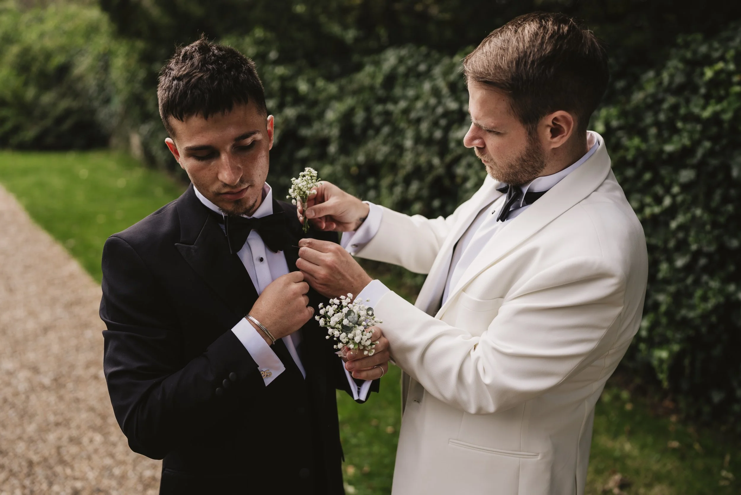 Two men in tuxedos, one in a black tuxedo and the other in a white tuxedo, outside with greenery, as the man in white adjusts the lapel or boutonniere of the man in black.