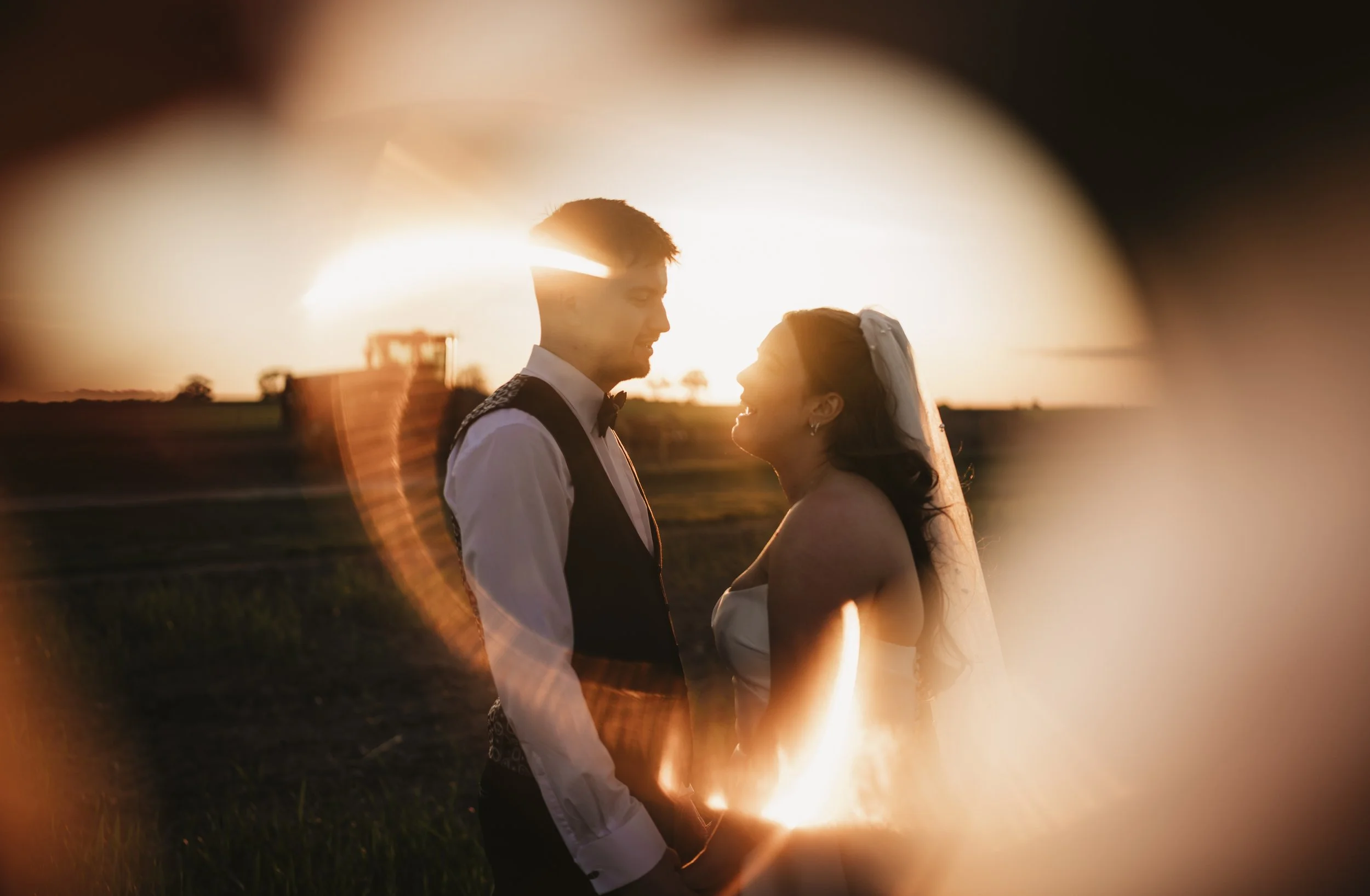 A couple dressed in wedding attire facing each other at sunset, with the photo taken through a circular lens or opening creating a warm glow and framing the scene.