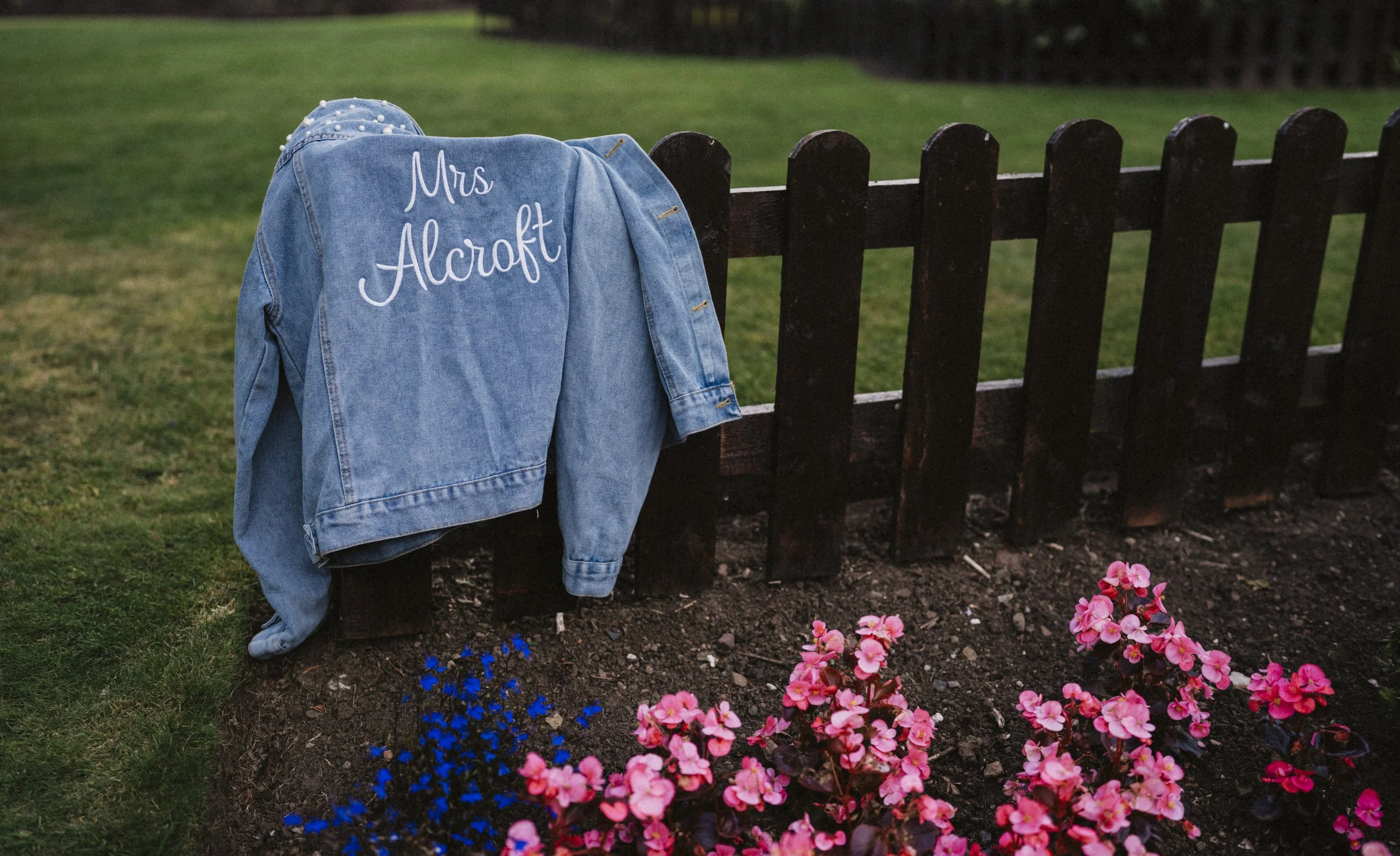 A denim jacket with 'Mrs Alcroft' embroidered on the back hanging on a dark wooden fence near a flower bed with pink and purple flowers, on a grassy lawn.