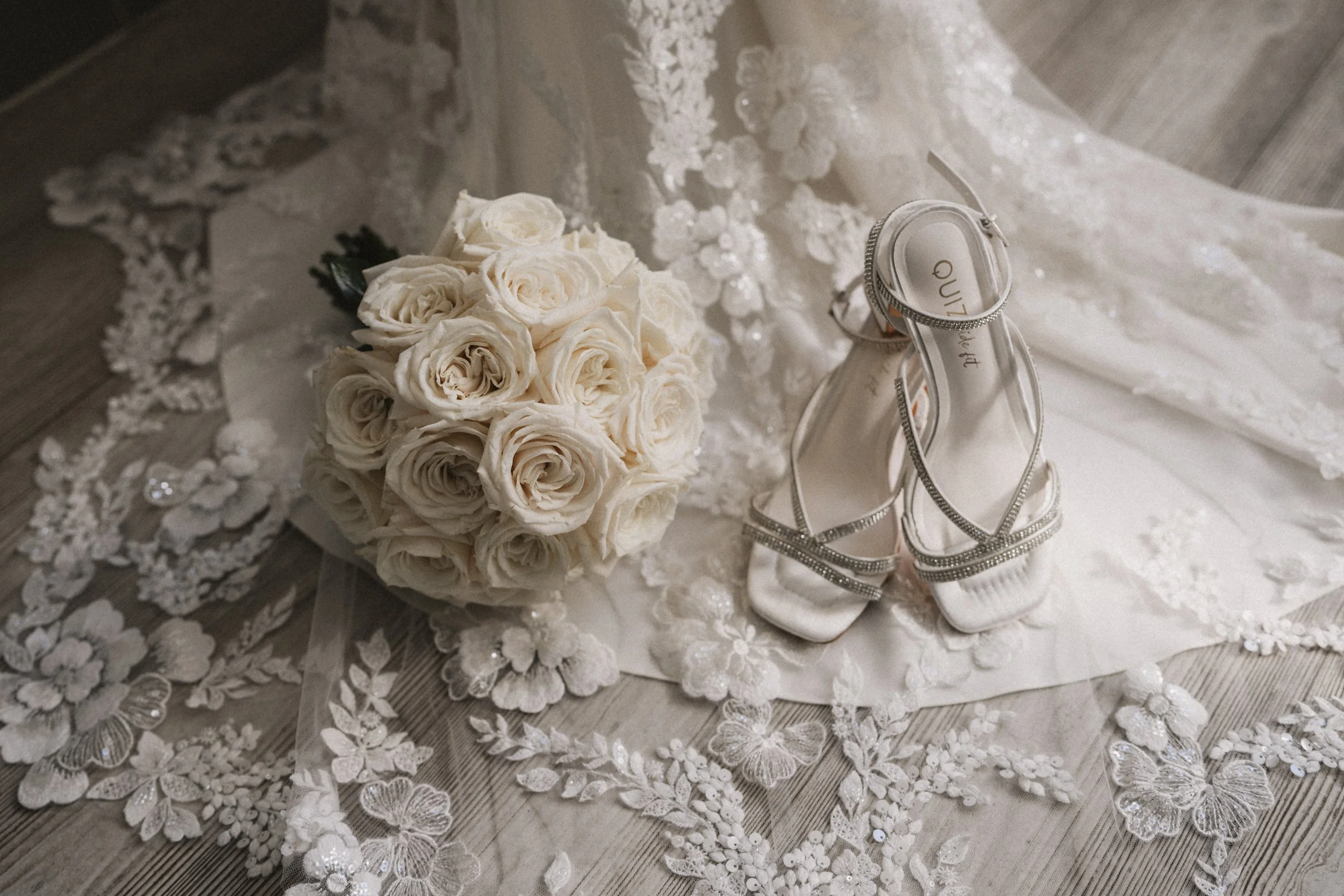 A bouquet of cream-colored roses, a pair of white high-heeled shoes with rhinestone straps, and an embroidered lace fabric are arranged on a wooden floor.