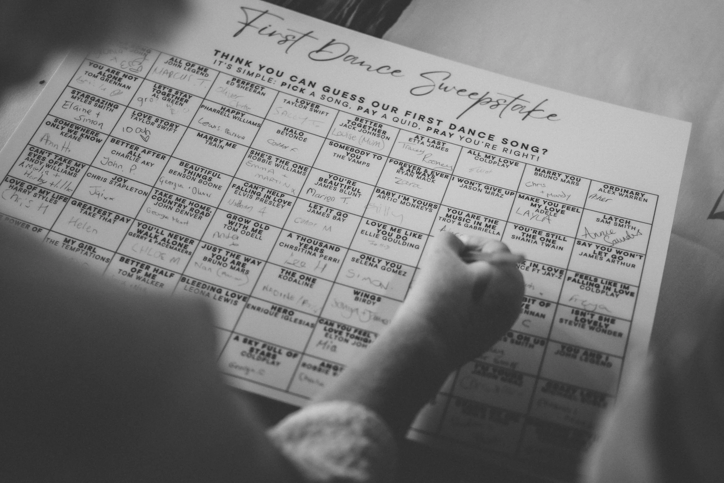 A person filling out a crossword puzzle titled 'First Dance Sweepstakes' with a pen. The puzzle features song titles under the heading 'Think you can guess our first dance song? It's simple: pick a song, pay a quid, pray you're right!' The crossword 