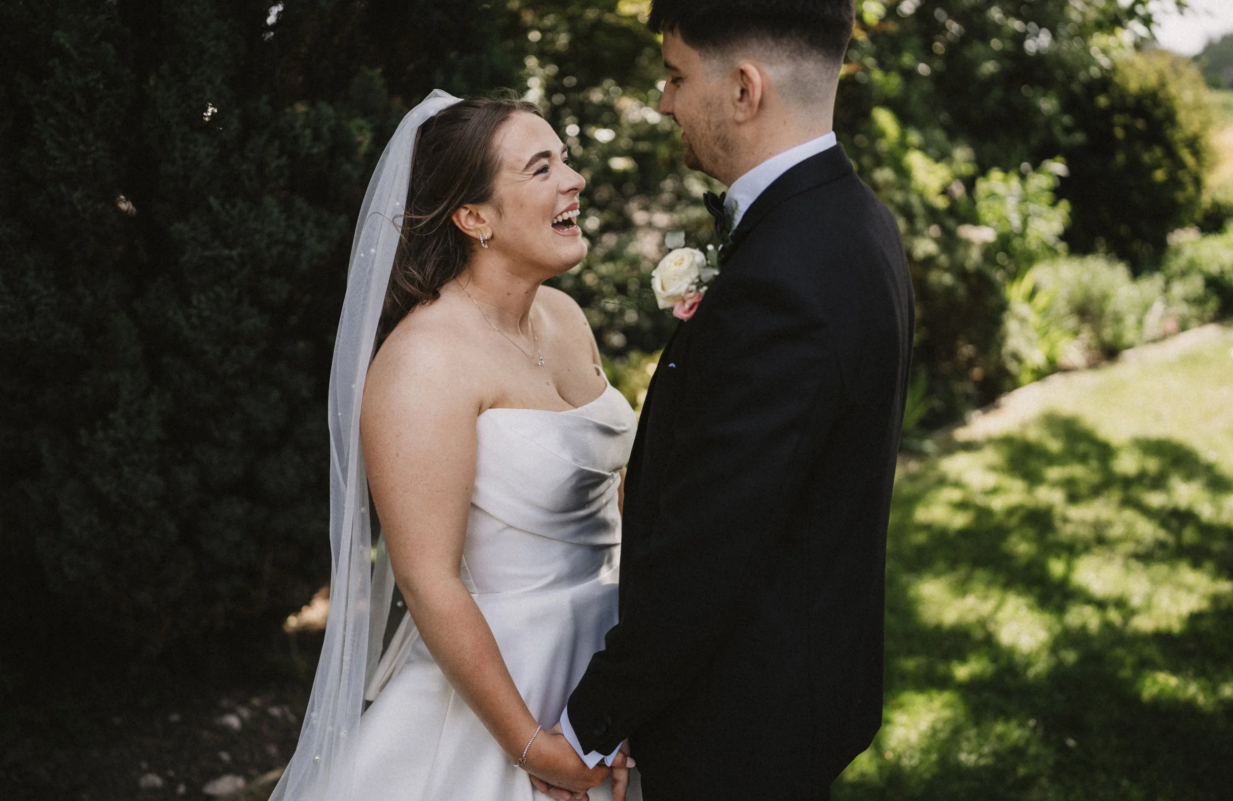 Bride and groom standing outdoors, holding hands, smiling, facing each other in wedding attire with greenery background.