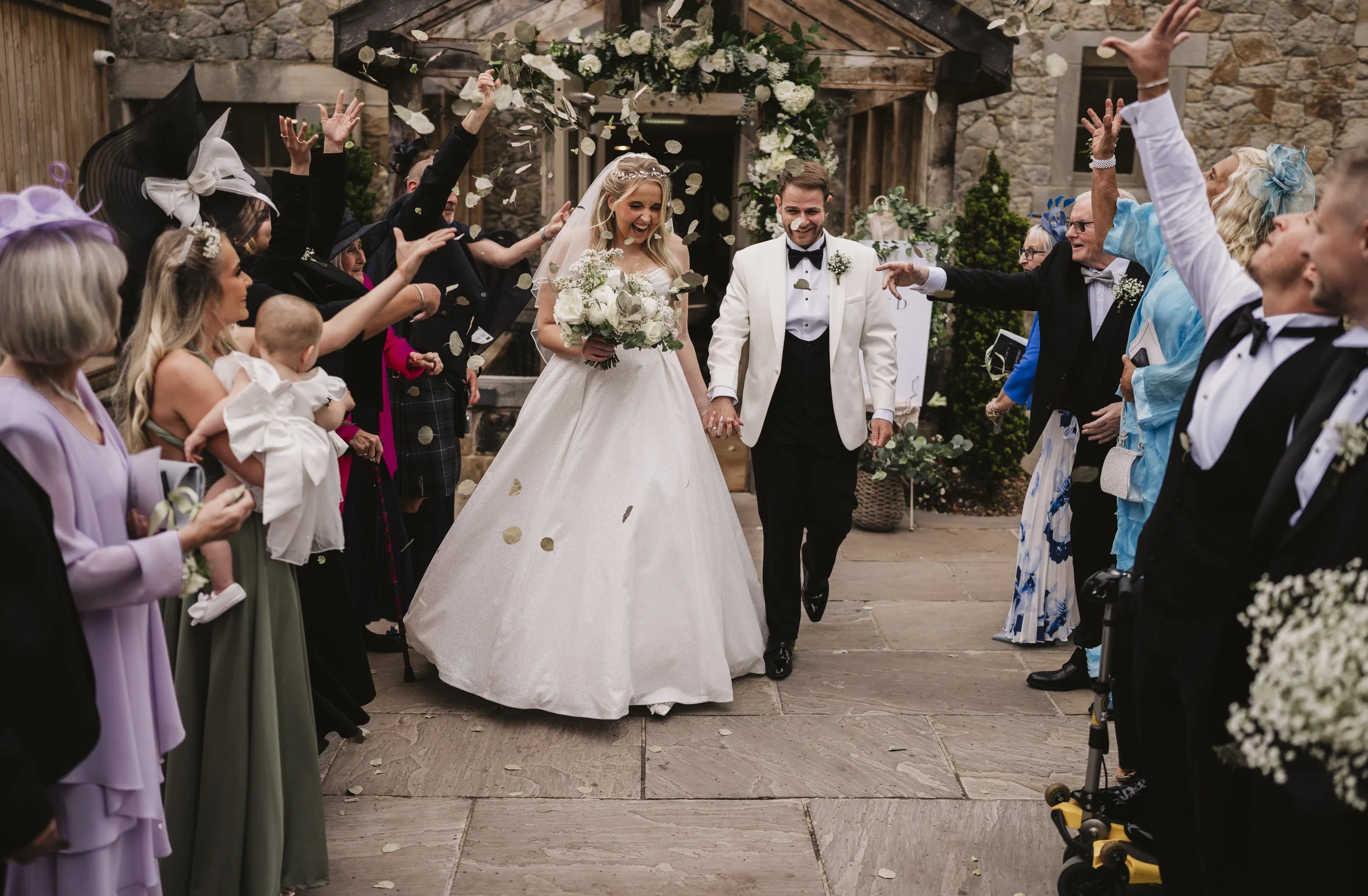 A bride and groom walking hand in hand surrounded by guests throwing flower petals in the air at their wedding celebration outdoors.