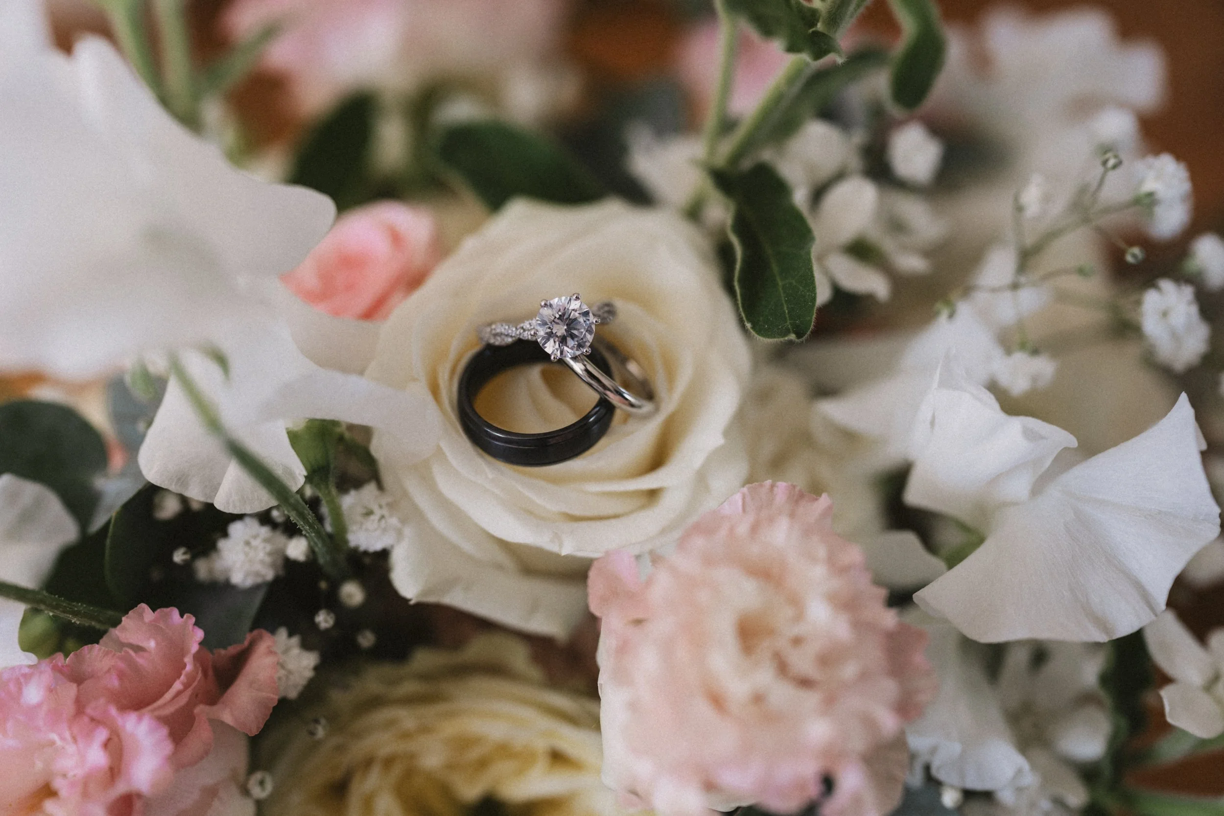 Close-up of an engagement ring with a large diamond and a wedding band placed on a white rose surrounded by other pink and white flowers and green leaves.