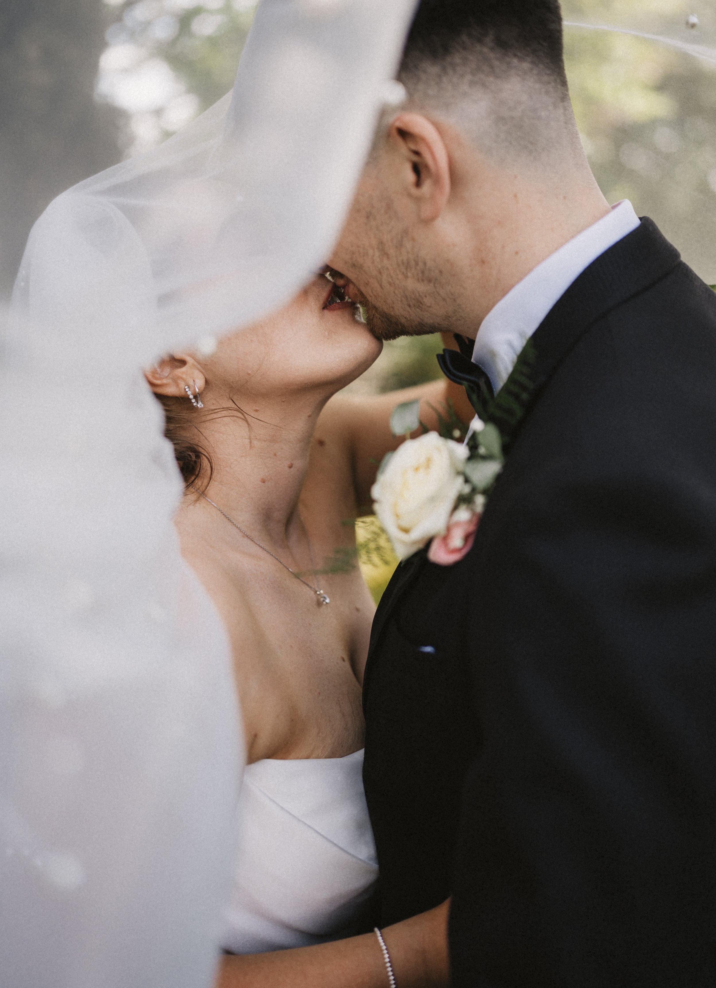 Close-up of a bride and groom kissing under a wedding veil outdoors, with the bride wearing a white strapless dress, necklace, and earrings, and the groom in a black tuxedo with a boutonnière.