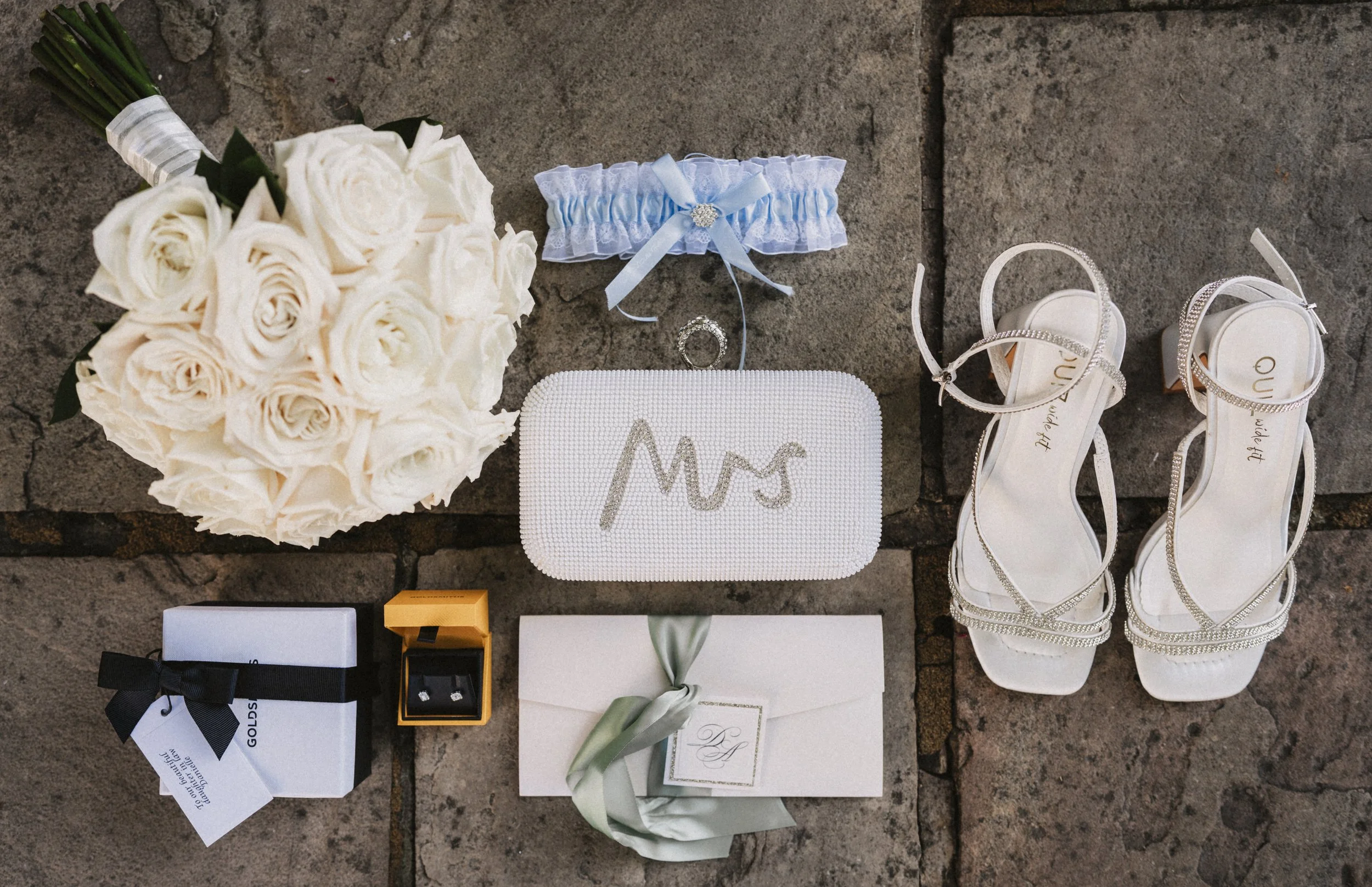 A wedding bouquet of white roses, a blue garter, a wedding ring, white high-heeled sandals, a white box with a black ribbon and tag, a small orange box with earrings, a white envelope with a green ribbon, and a white clutch with "Mrs" written on it, 