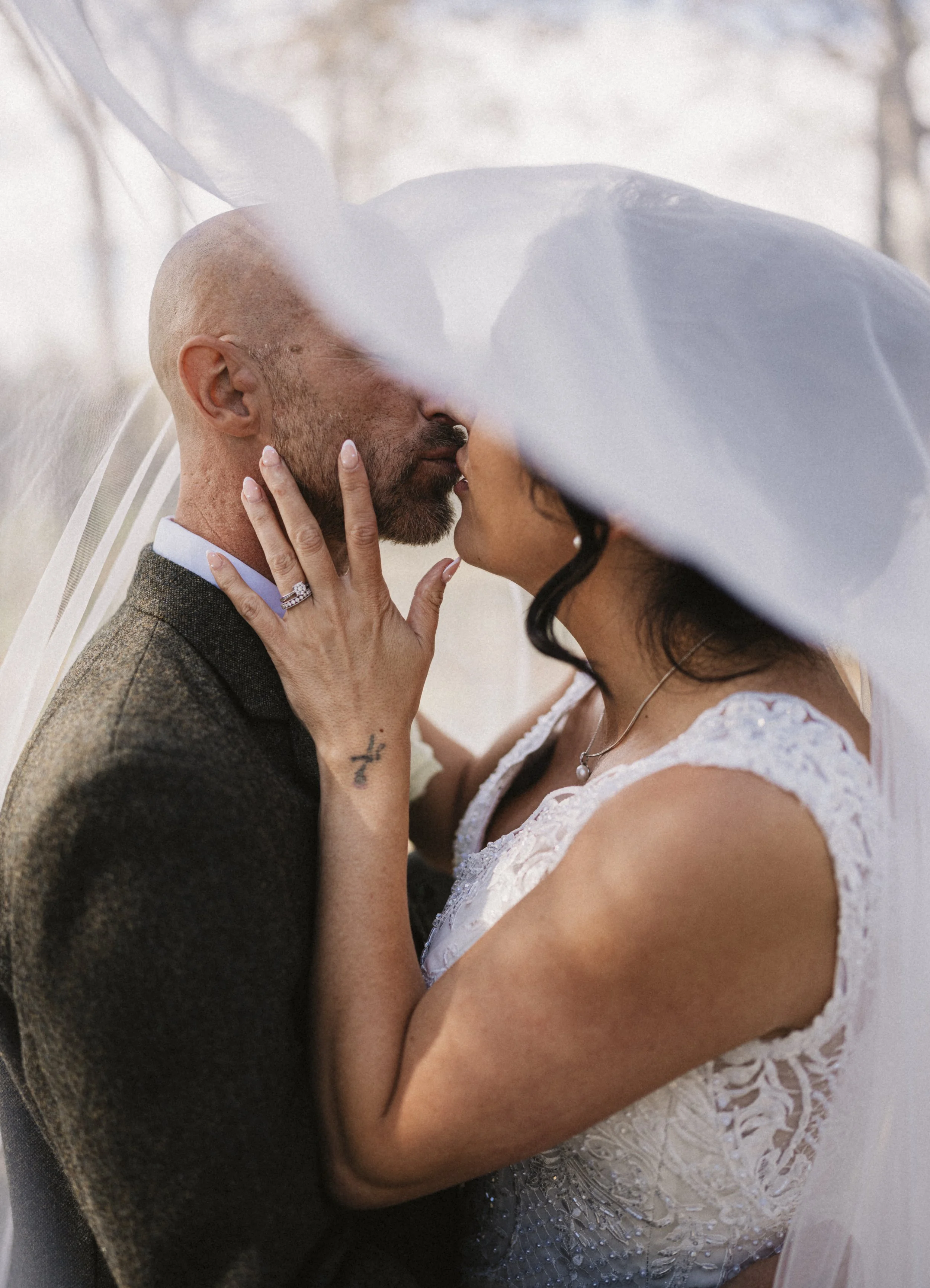 A wedding couple sharing a kiss under a veil outdoors, with the bride's hand on the groom's face, the bride wearing a white lace dress and a wedding band, and the groom in a brown tweed jacket.