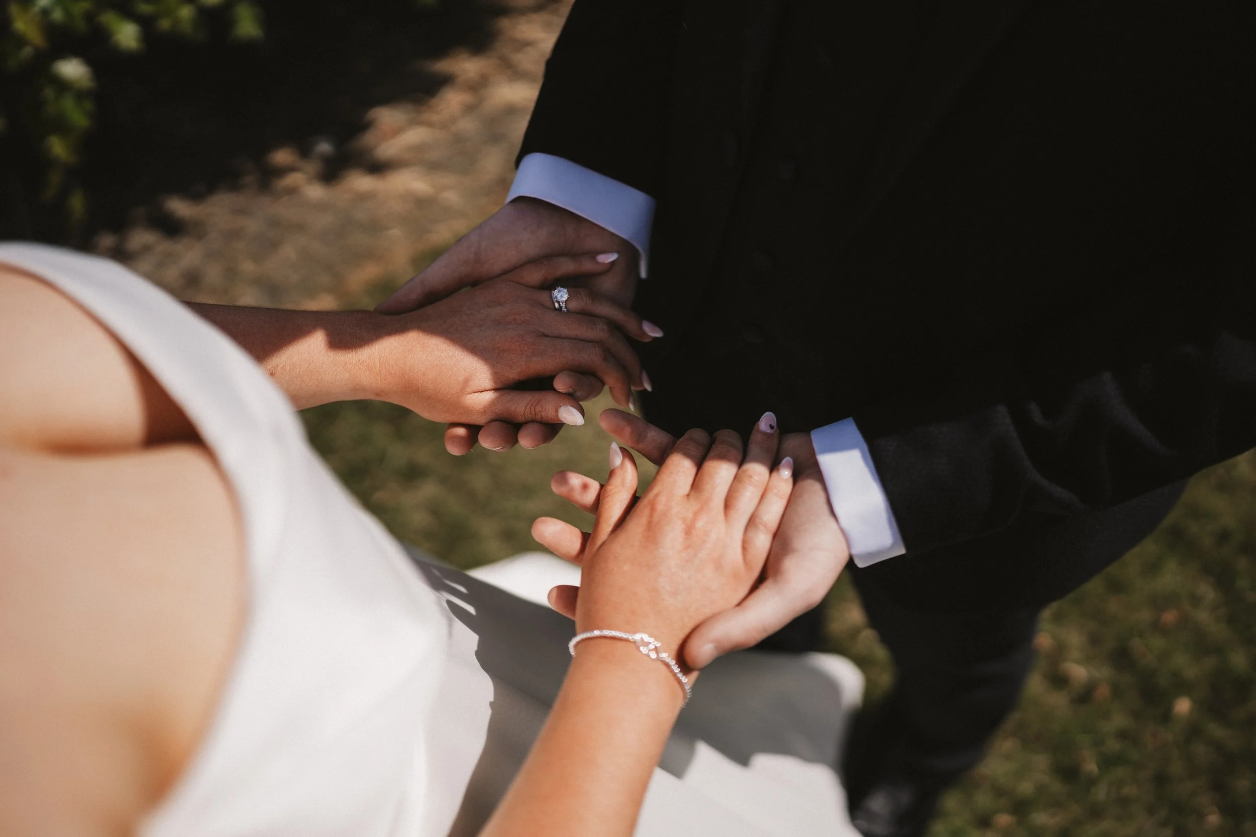 A bride and groom holding hands during their wedding ceremony, with the bride wearing a white dress and jewelry, and the groom in a black suit with white cuffs.