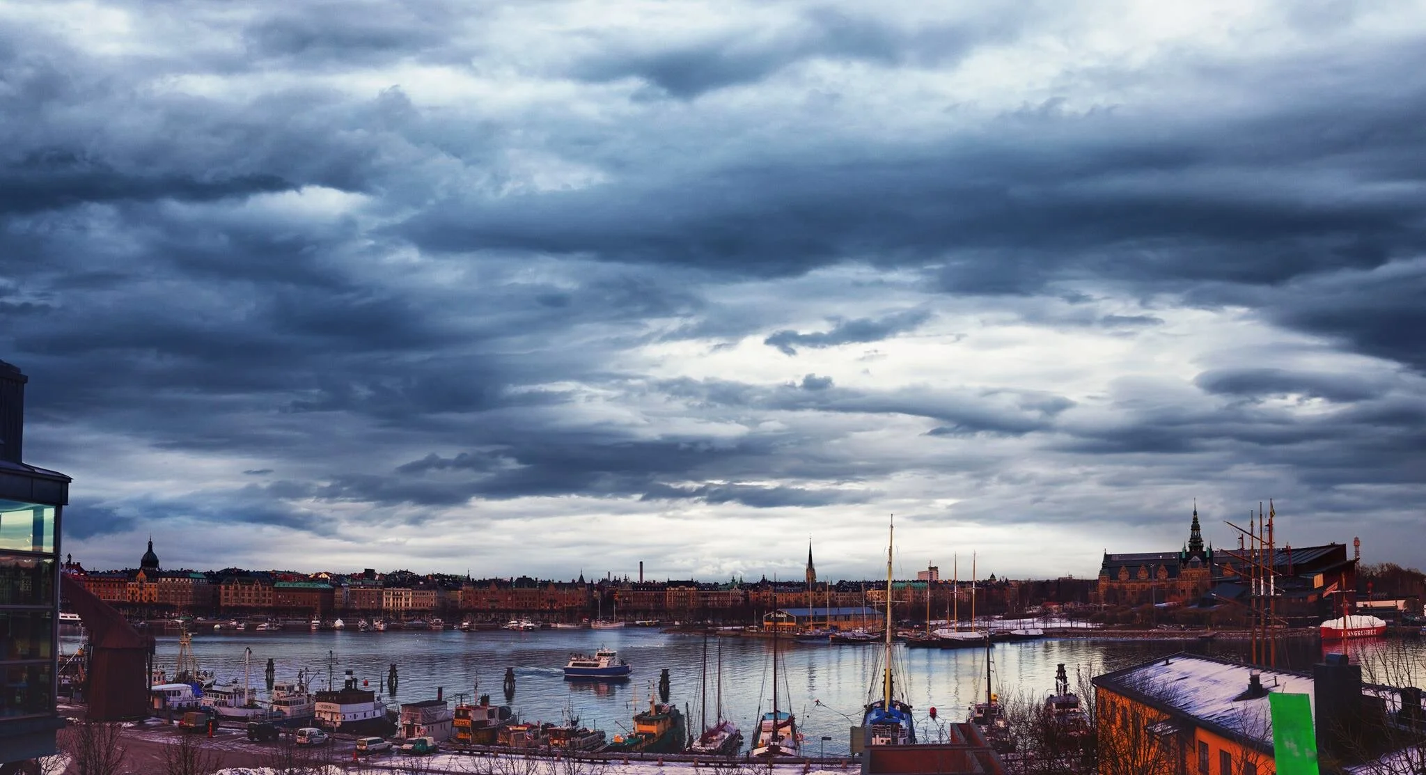 Overlooking a harbor with boats under a cloudy sky, with historic buildings and church spires in the background.