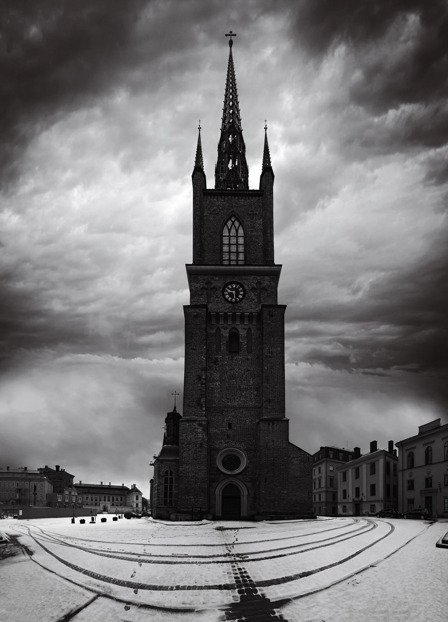 A tall, gothic-style church tower with a clock, set against a dark, cloudy sky, with snow-covered ground and footprints leading to the entrance.