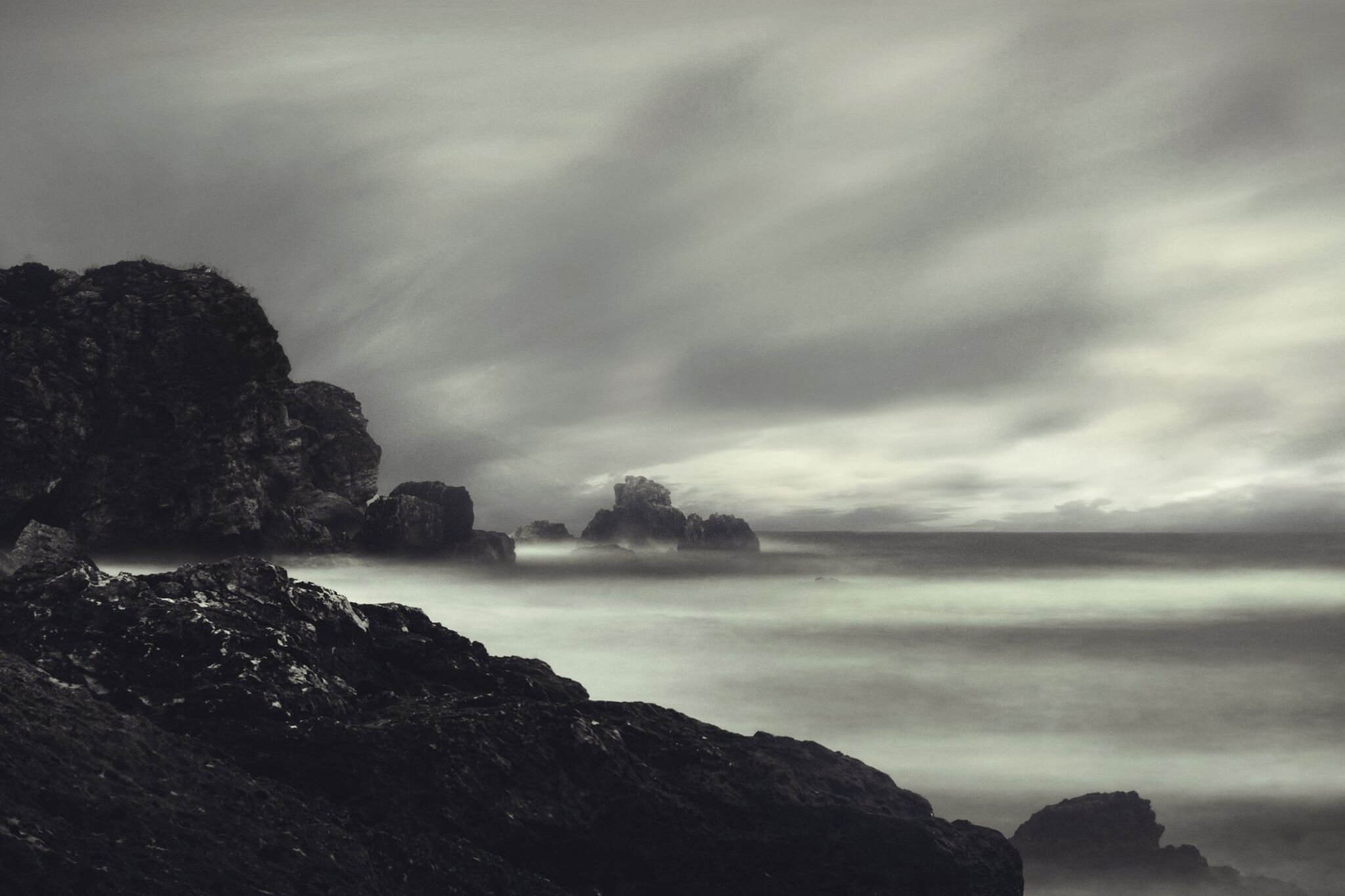 Dark rocky shoreline with large rocks near the coast, under a cloudy, overcast sky, and waves washing over the rocks in a long exposure shot.