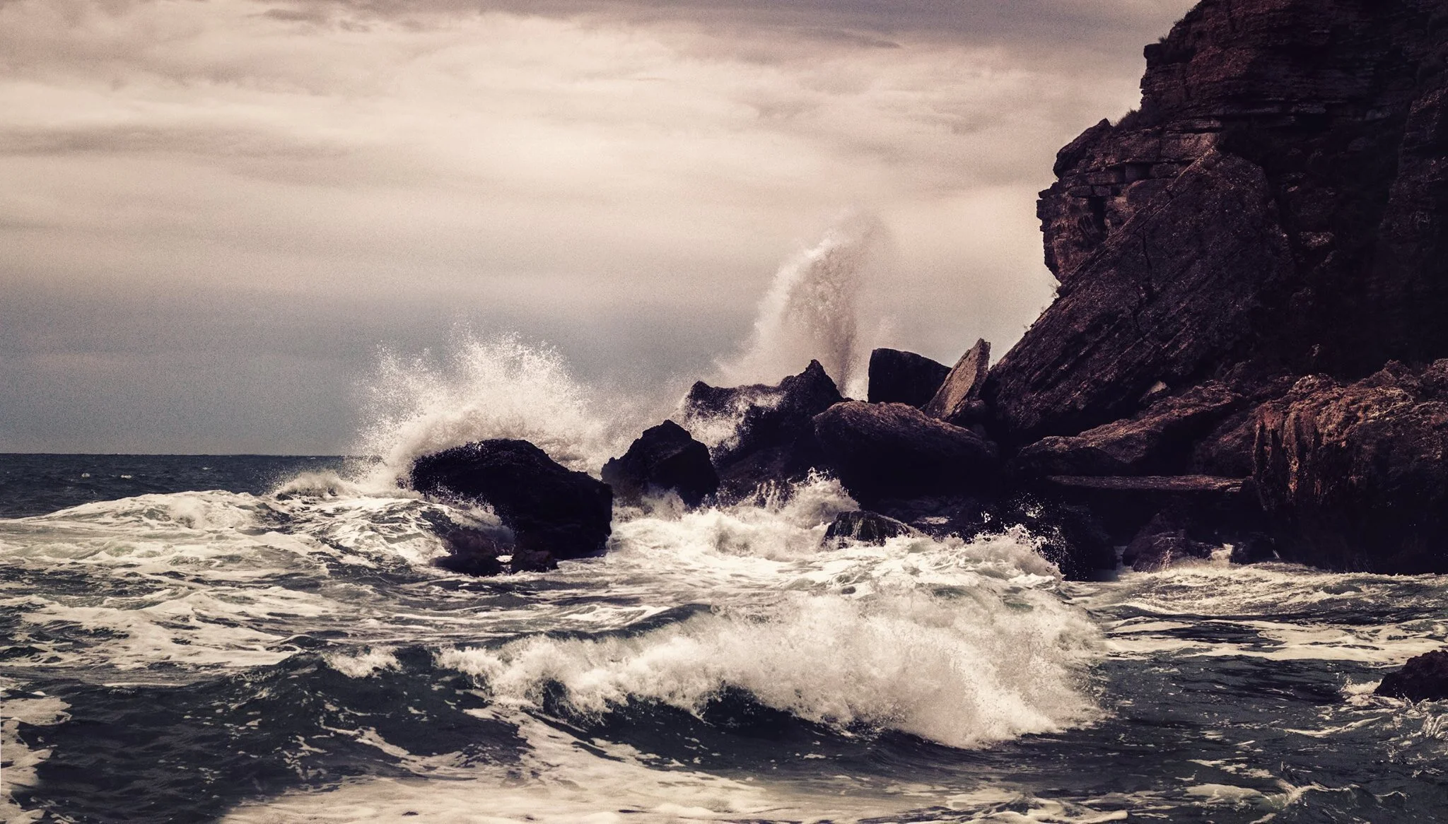 Dark ocean waves crashing against rocks near a steep cliff, under a cloudy sky.