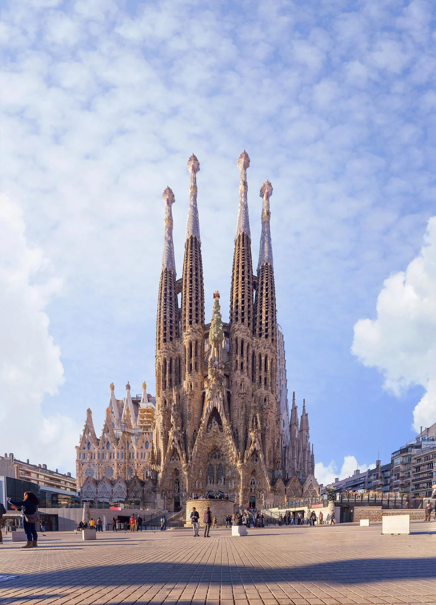 Sagrada Família basilica with tall spires and detailed facade in Barcelona, Spain, under a blue sky with clouds, with people walking on the plaza in front.
