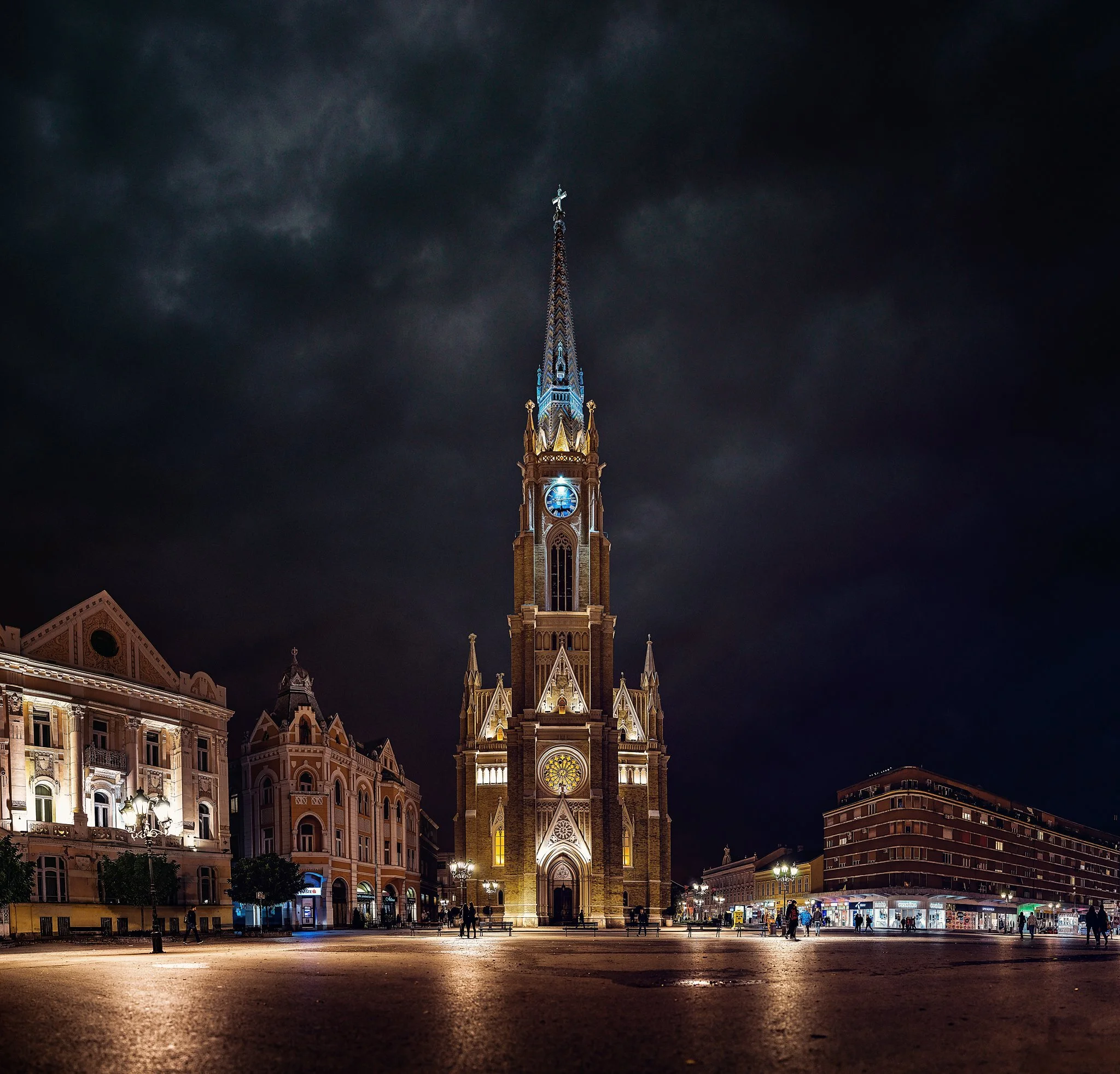 Night view of a tall illuminated church with a clock and pointed steeple in a city square, surrounded by historic buildings and some people walking.