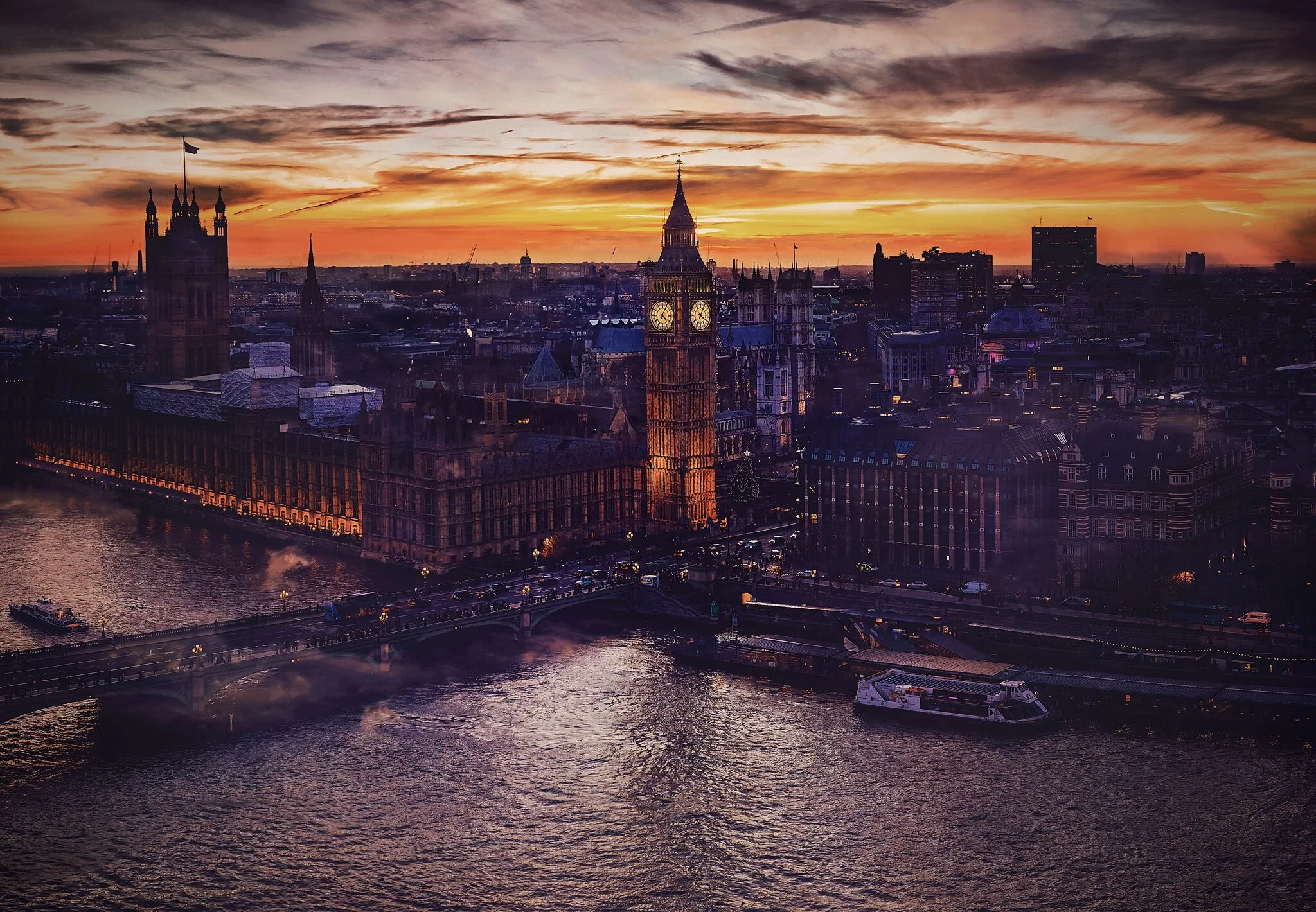 Sunset over the London skyline with Big Ben and the Houses of Parliament next to the River Thames, with boats and traffic on the bridge.